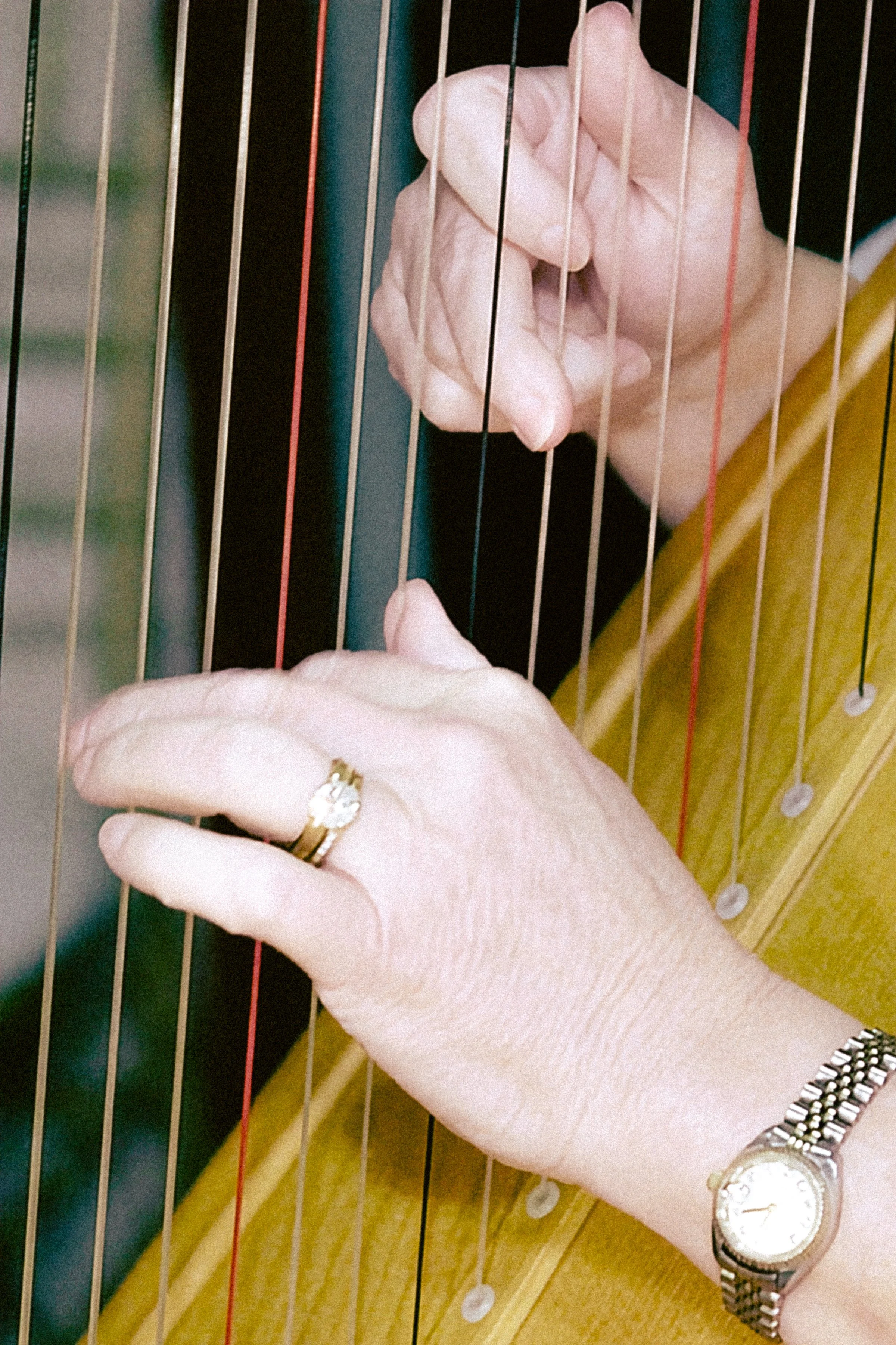 A person's hand with a silver watch and ring, holding the strings of a musical instrument, possibly a harp or similar stringed instrument, with metal and wooden components visible.