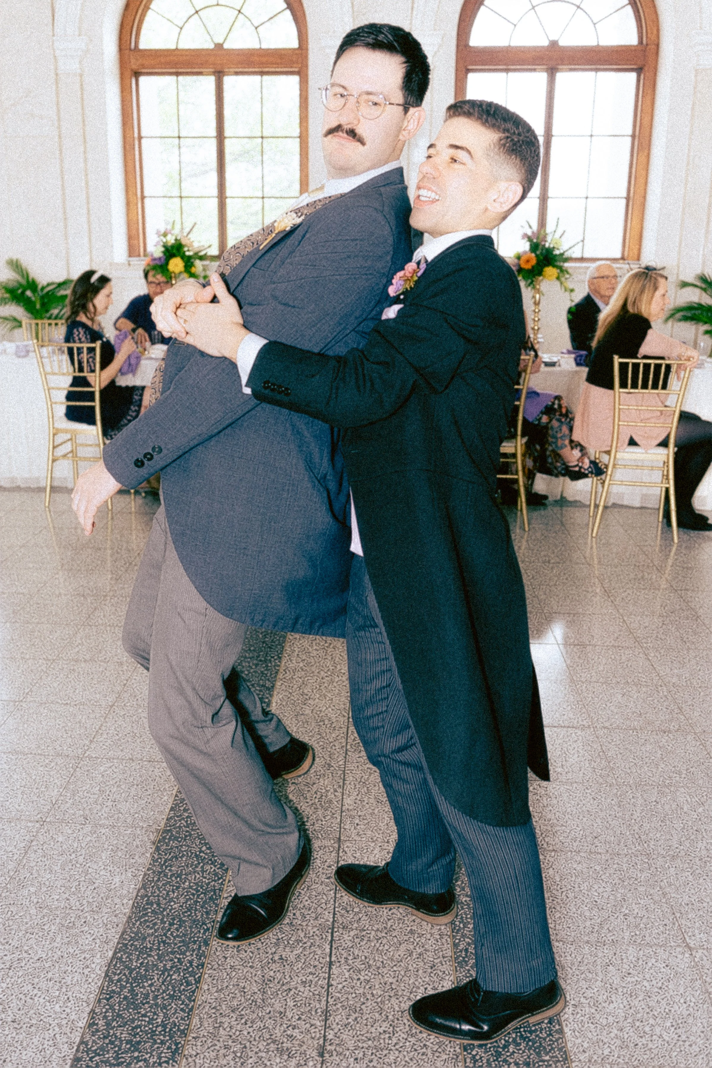 Two men dancing closely at a formal event in a bright room with large arched windows, surrounded by guests seated at tables with floral centerpieces.
