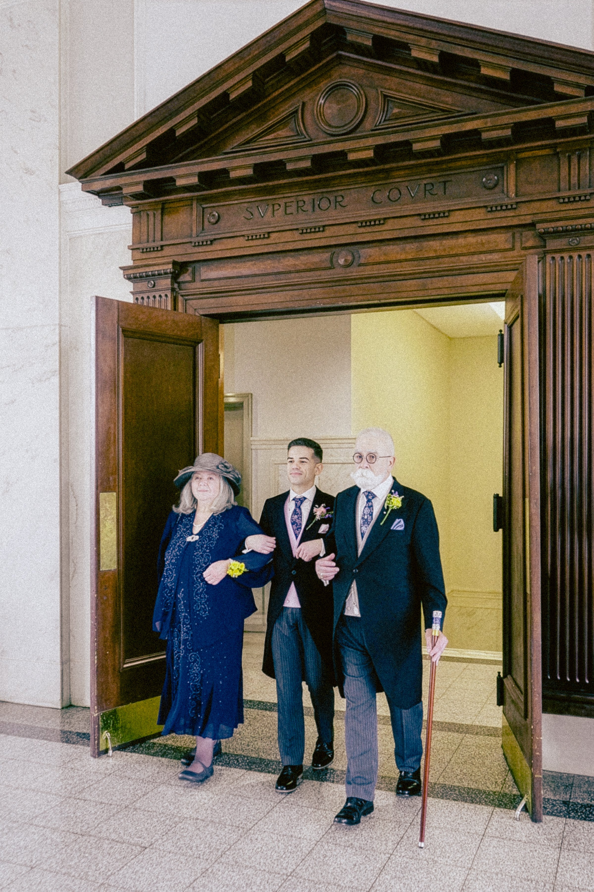 Three people walking through a large doorway with the text 'Superion Court' carved above it. The individuals are dressed in formal attire, and the setting appears to be inside a government or courthouse building.