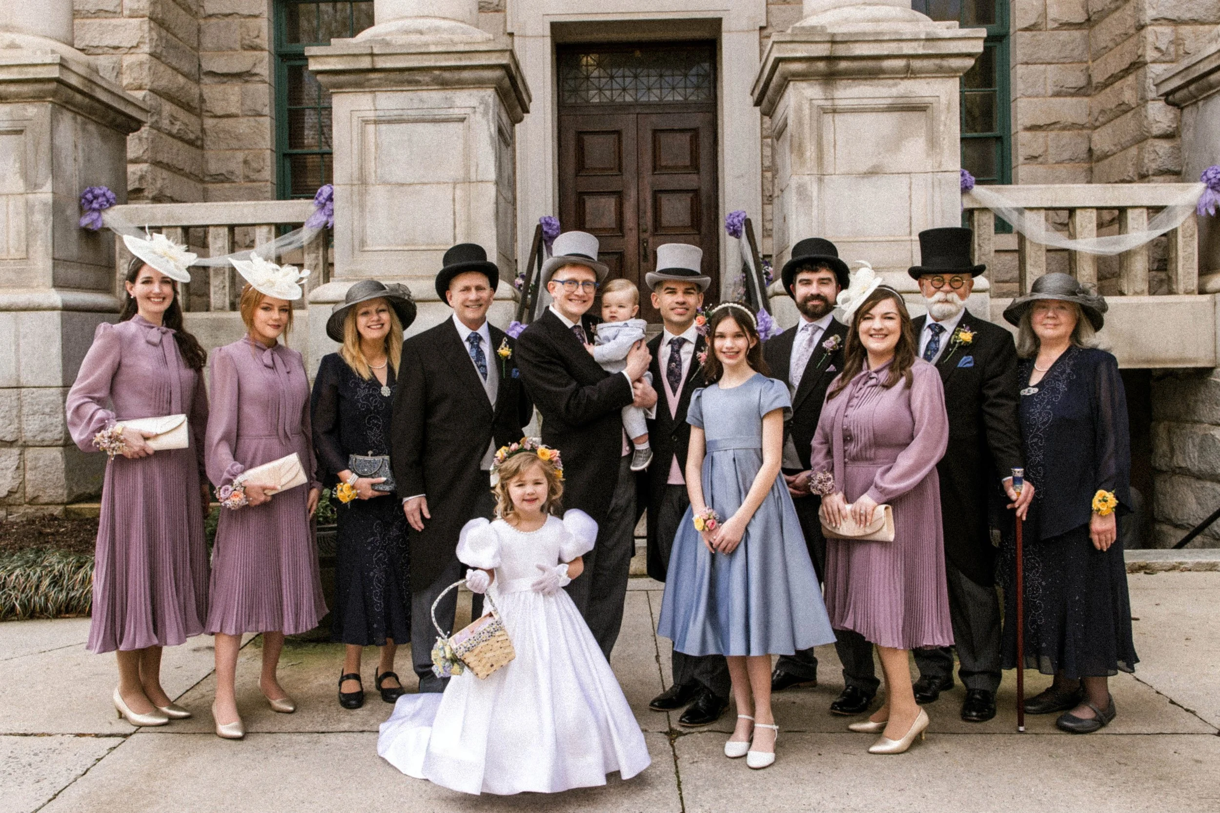 A group of people dressed in vintage, formal attire standing on a step in front of a stone building, celebrating a wedding. The group includes women in lavender and black dresses wearing hats, men in suits with top hats, and children in dress clothin