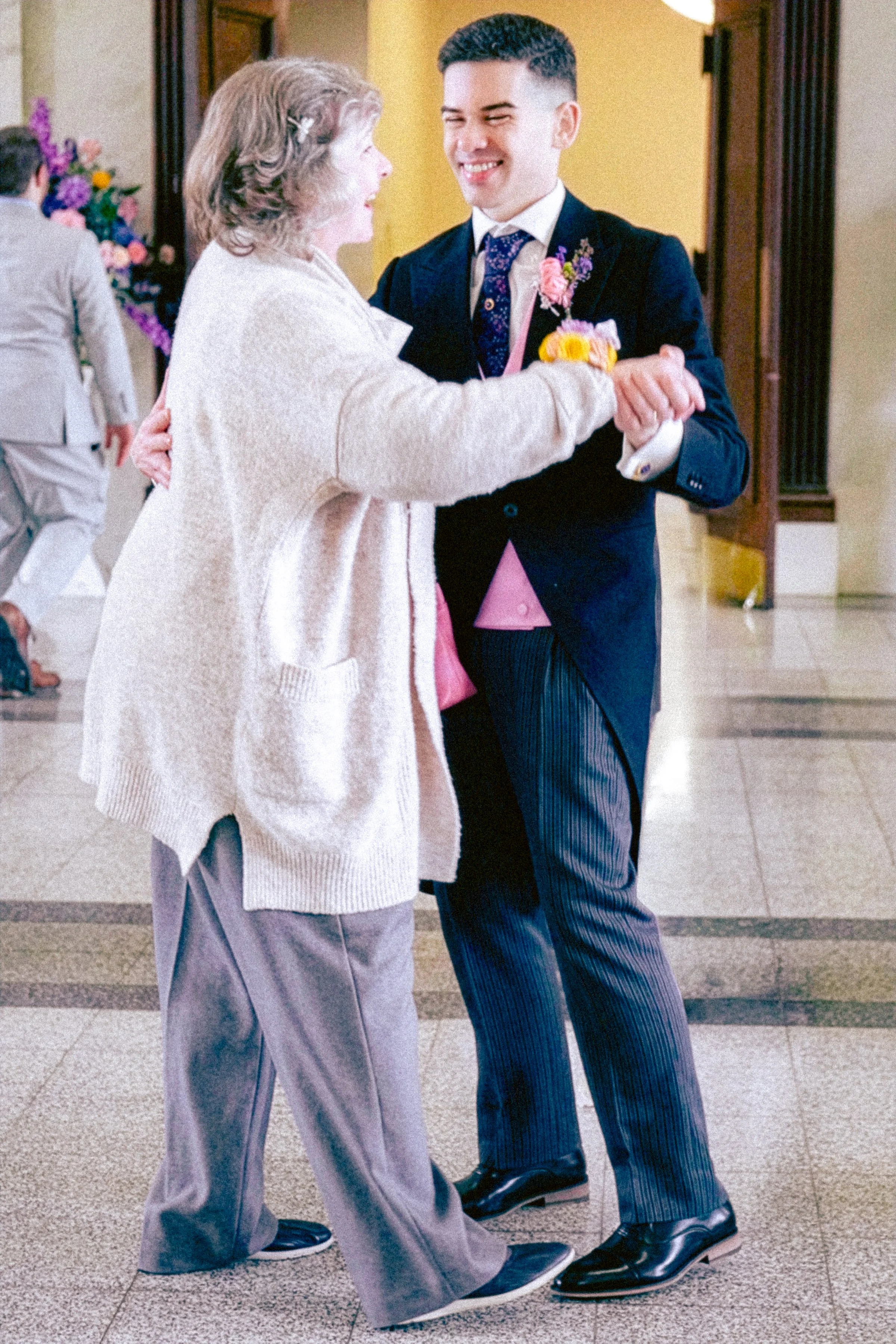 A young man in a dark suit with striped pants and a pink tie, smiling and dancing with an older woman in a light-colored coat, purple pants, and dark shoes, in an indoor setting with other people and colorful decorations in the background.