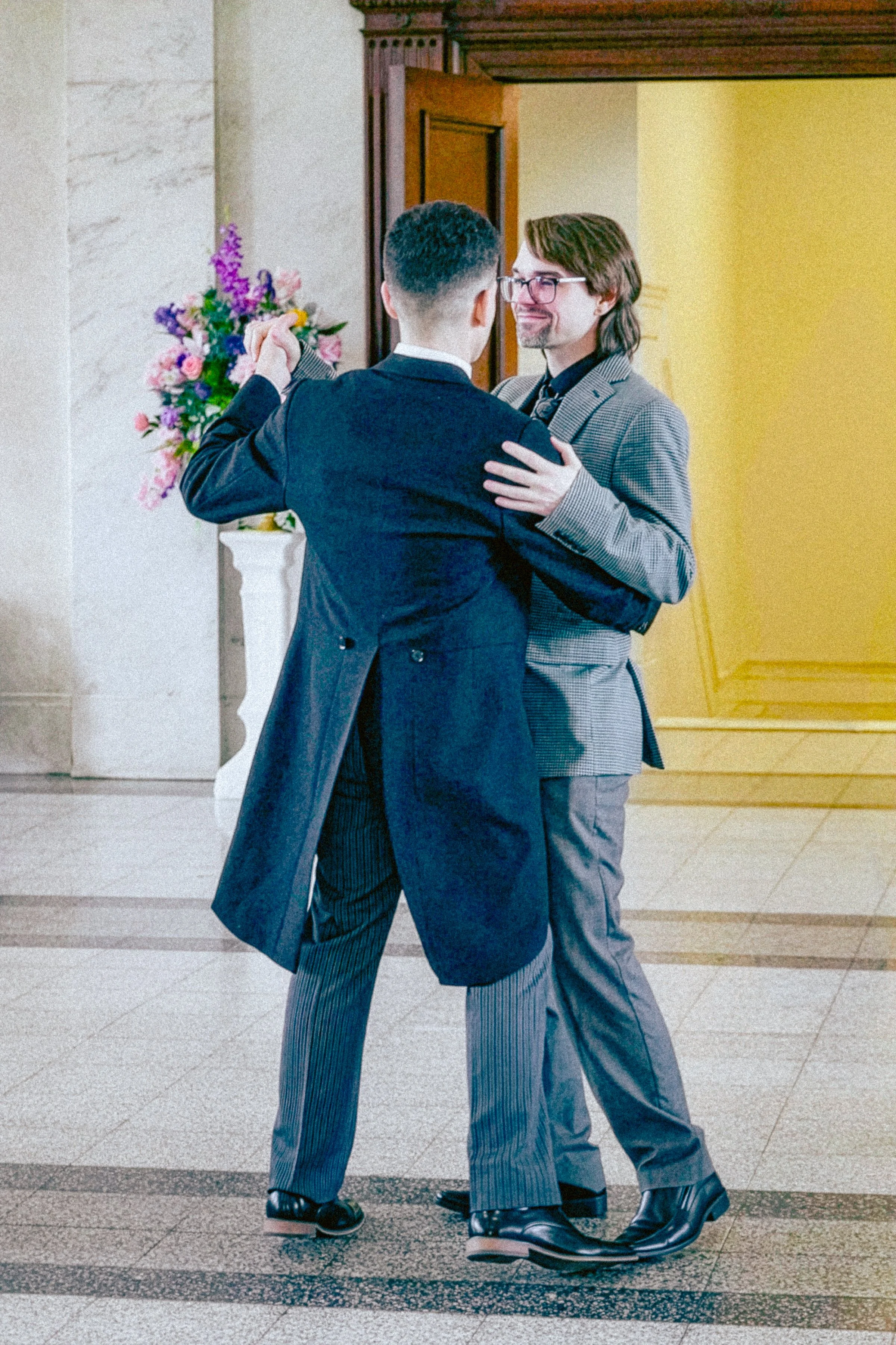 Two men dancing closely, holding each other, in an indoor setting with a vase of pink and purple flowers on a white pedestal in the background.