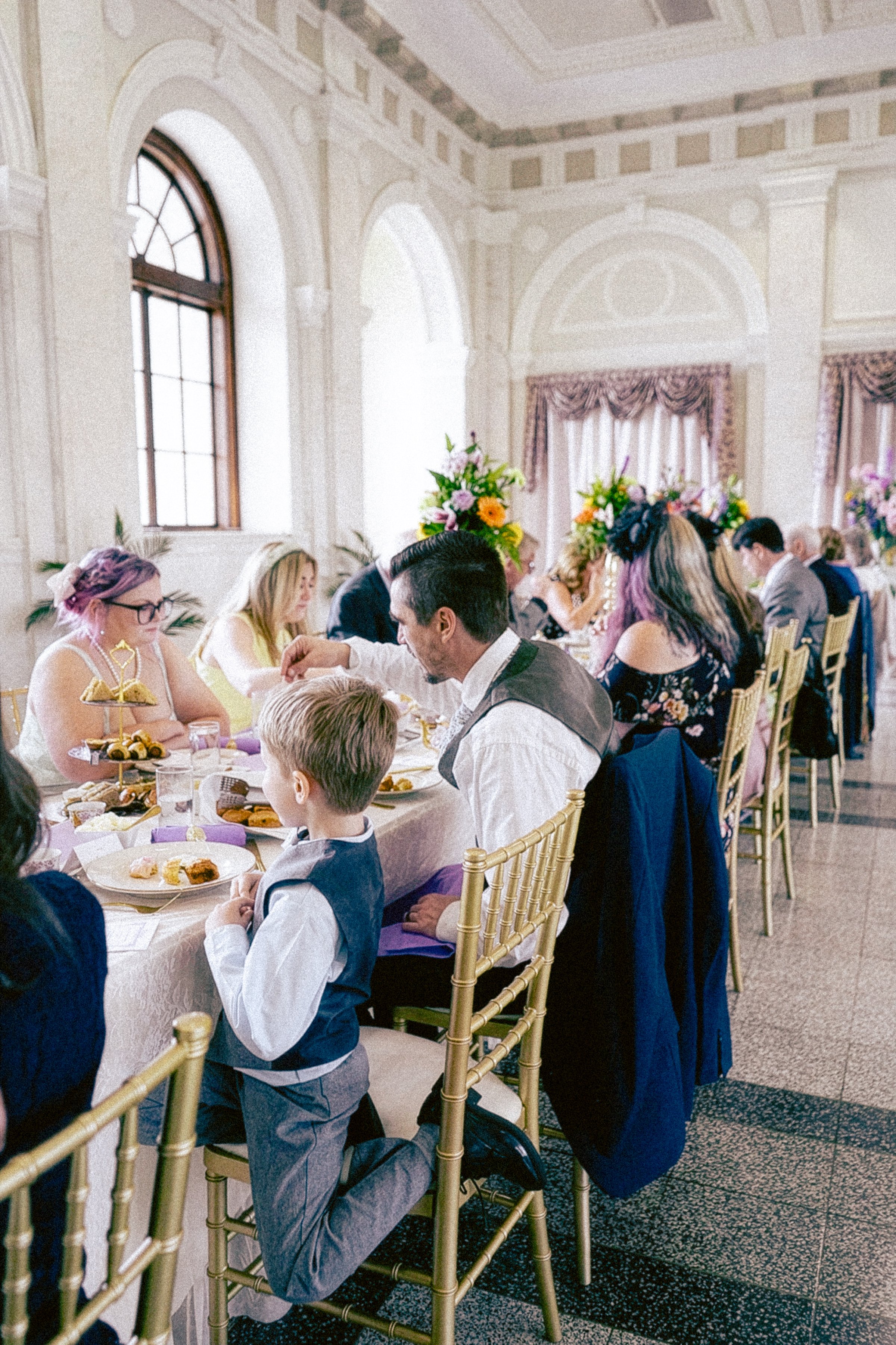 People sitting at a decorated wedding reception table with floral arrangements in an elegant room with large arched windows and ornate ceiling details.