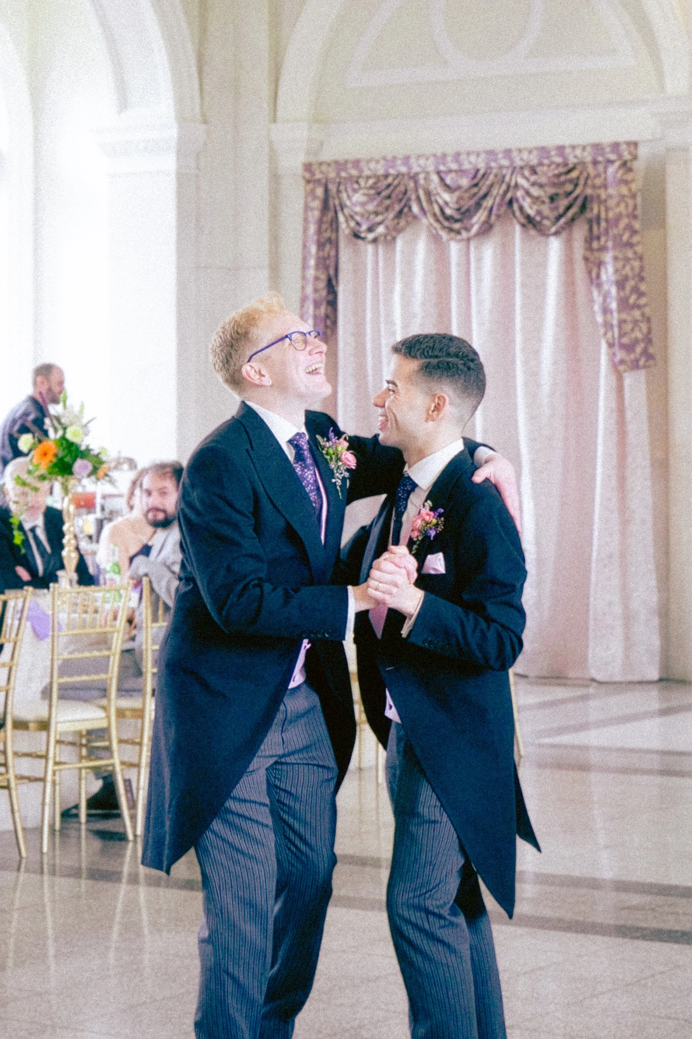 Two men in formal suits dancing together at a wedding reception, smiling and holding hands.