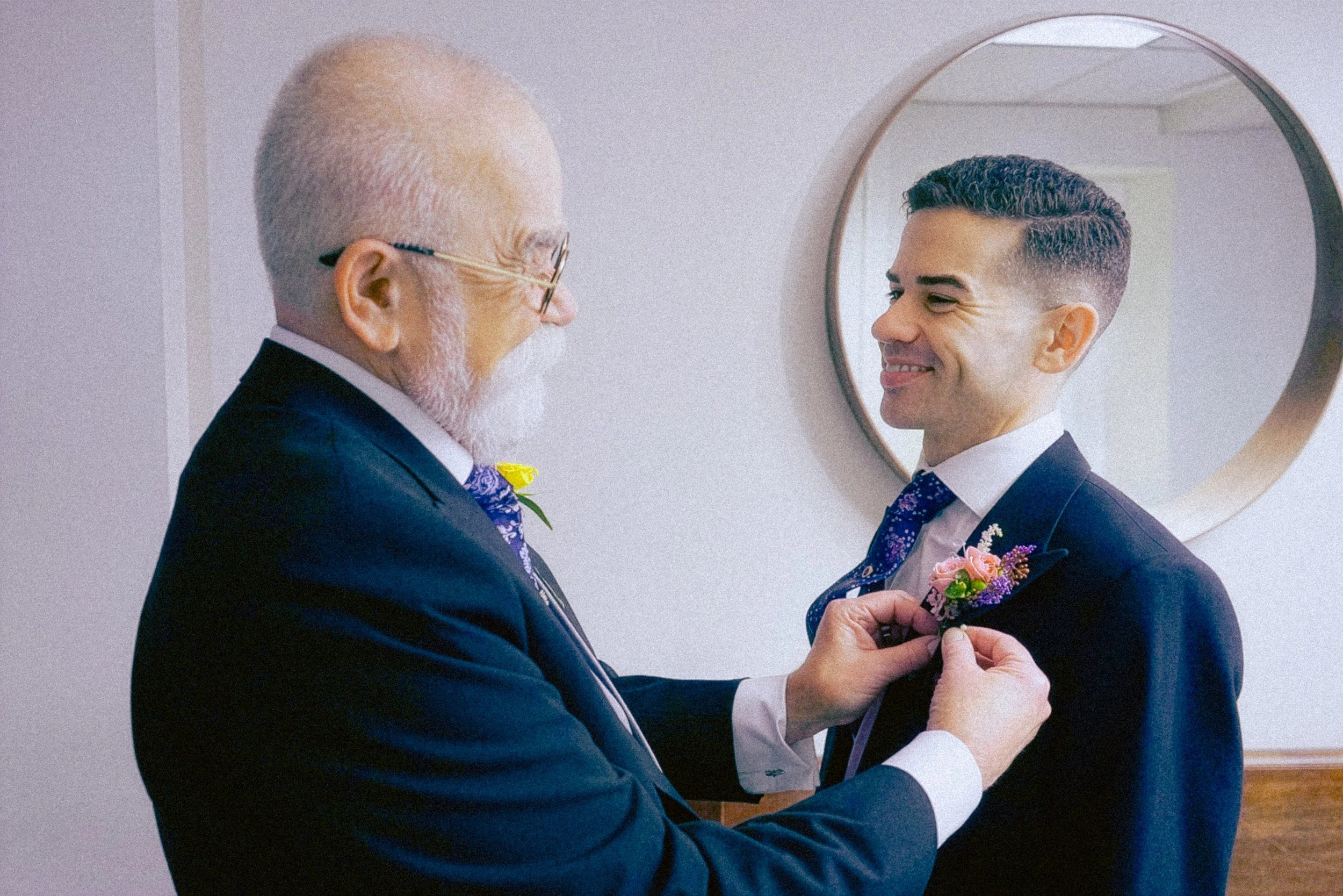 An elderly man with glasses and a white beard, wearing a dark suit and tie, is smiling as he helps a young man adjust his boutonniere in front of a mirror.