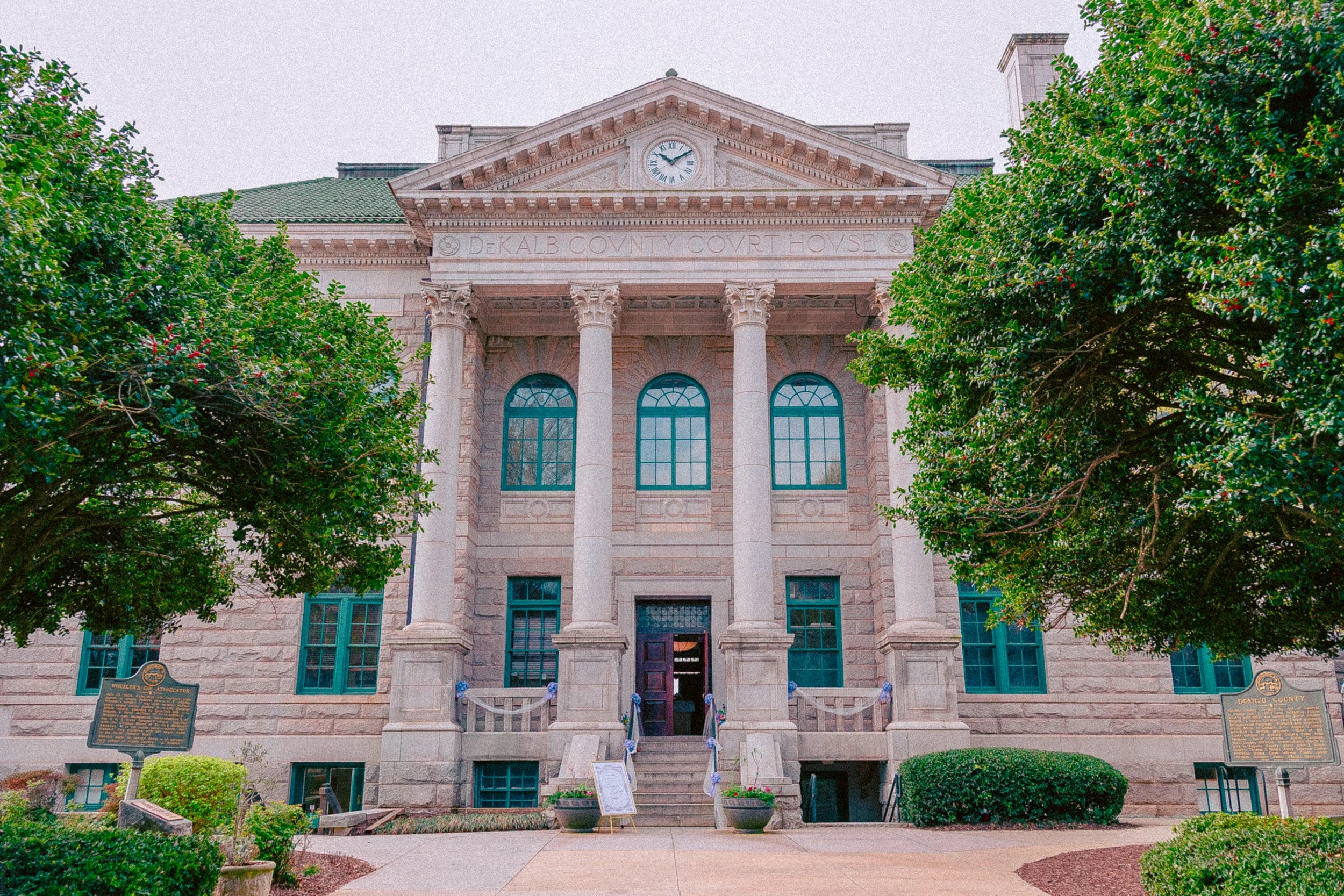 Front view of the Dekalb County Courthouse with a classical architectural style, featuring large columns, a clock at the pediment, and trees on either side.