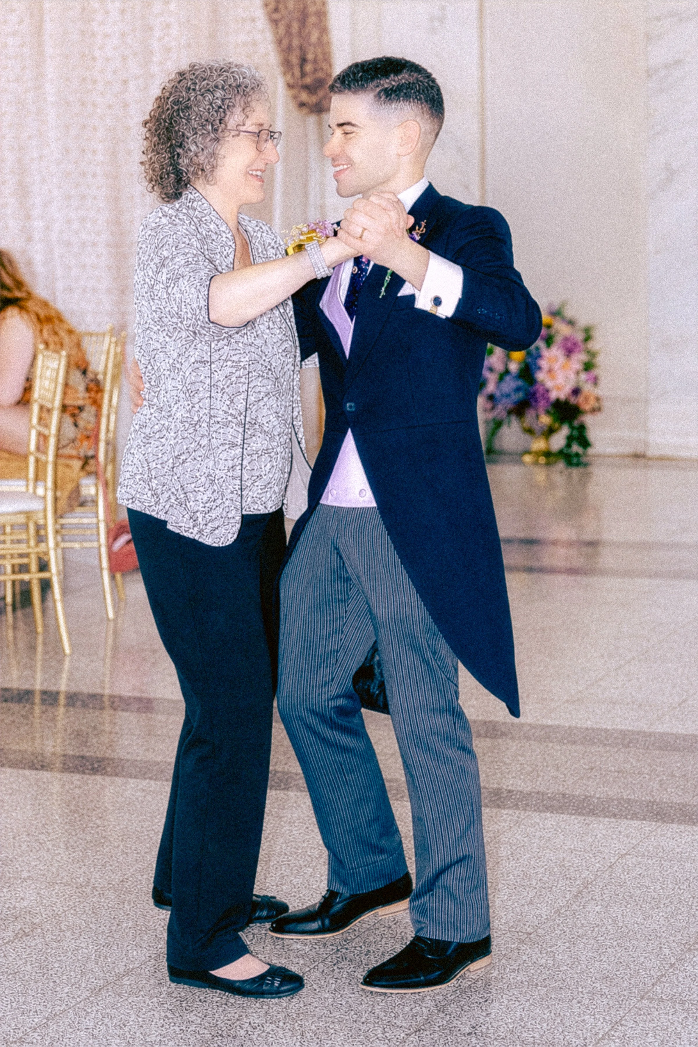 A couple dancing together at a formal event, holding hands and smiling, with a woman wearing glasses and a patterned blouse, and a man in a dark suit with striped pants.