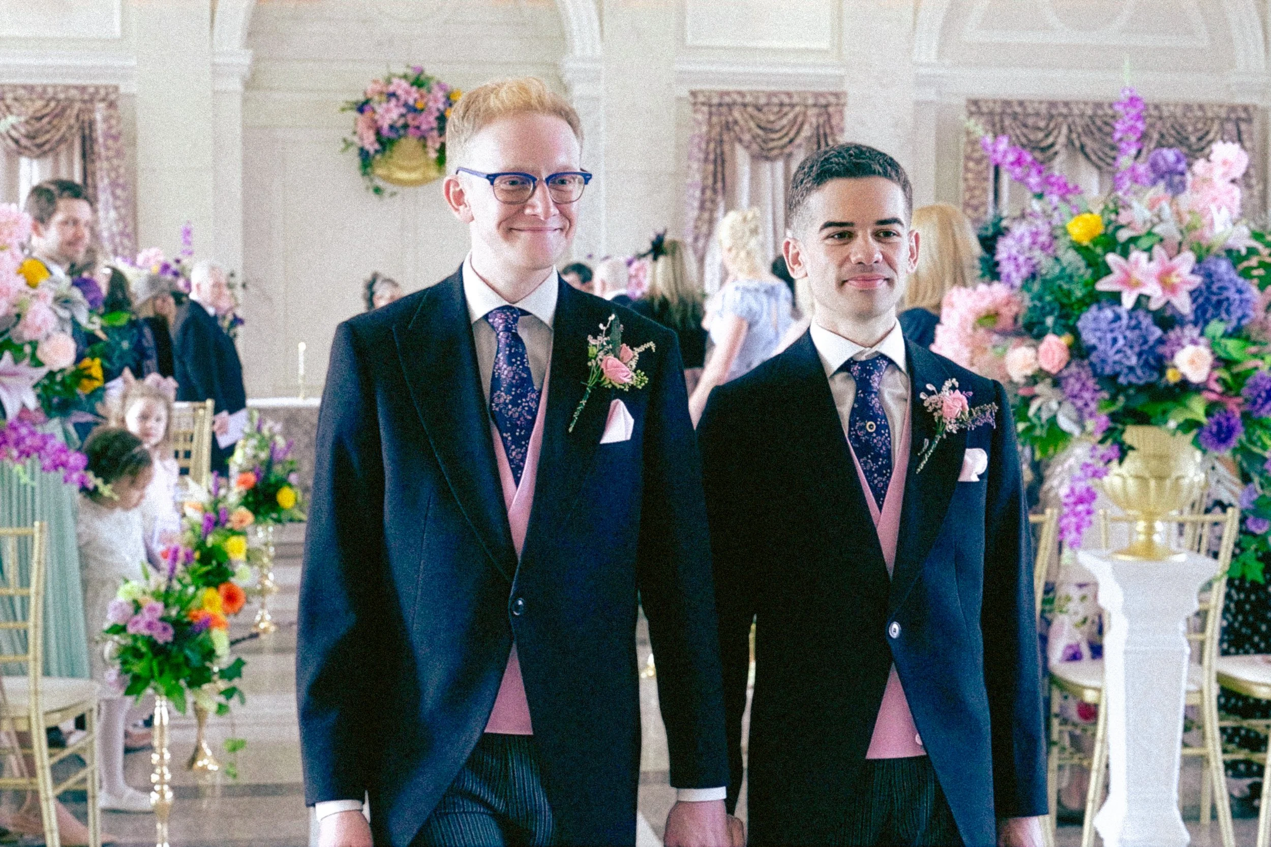 Two men dressed in suits with pink accents, standing in a decorated banquet hall with flowers, during a wedding reception.