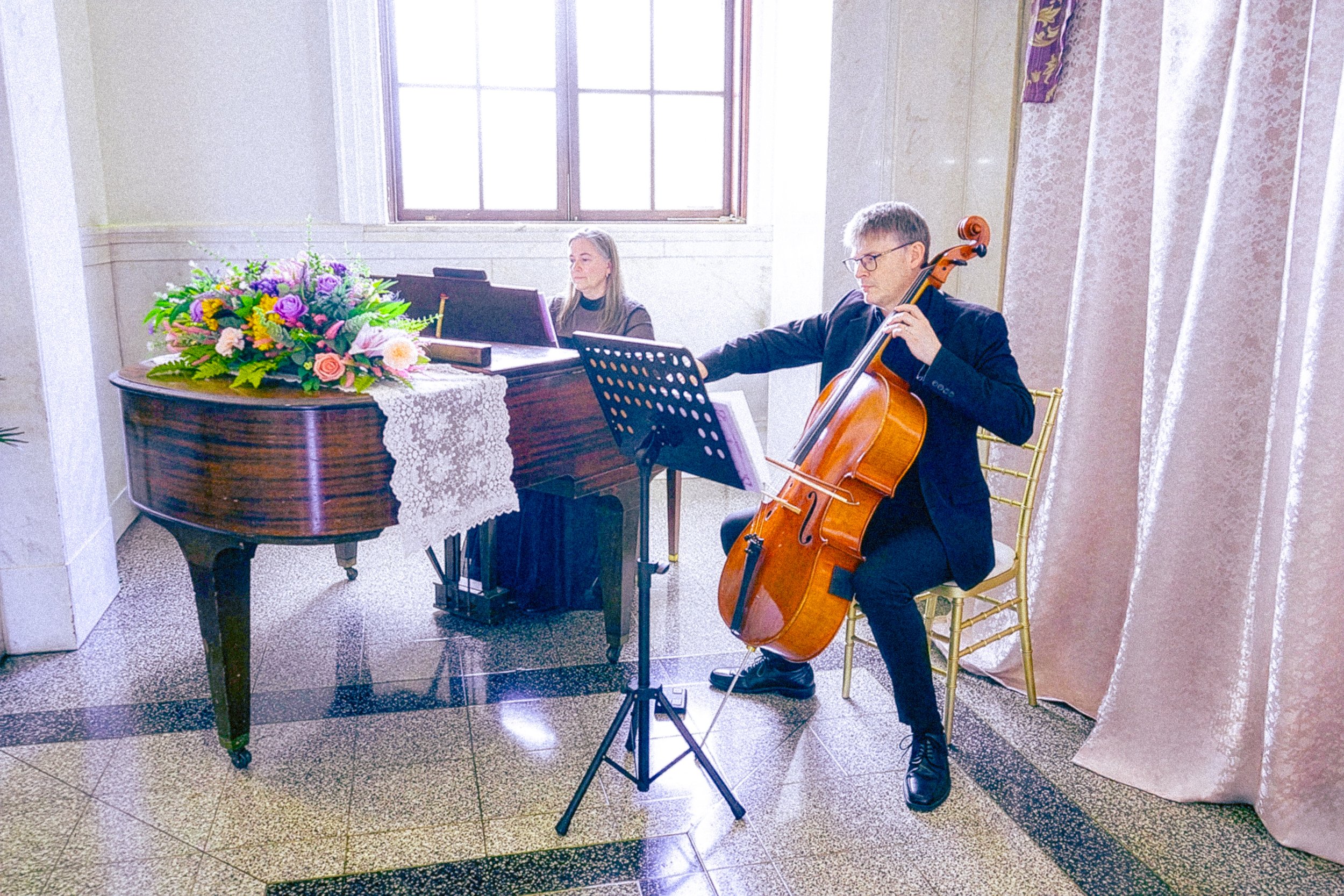 A man playing a cello while a woman plays a piano in a well-lit room with large window, floral arrangement on piano, and elegant curtains.