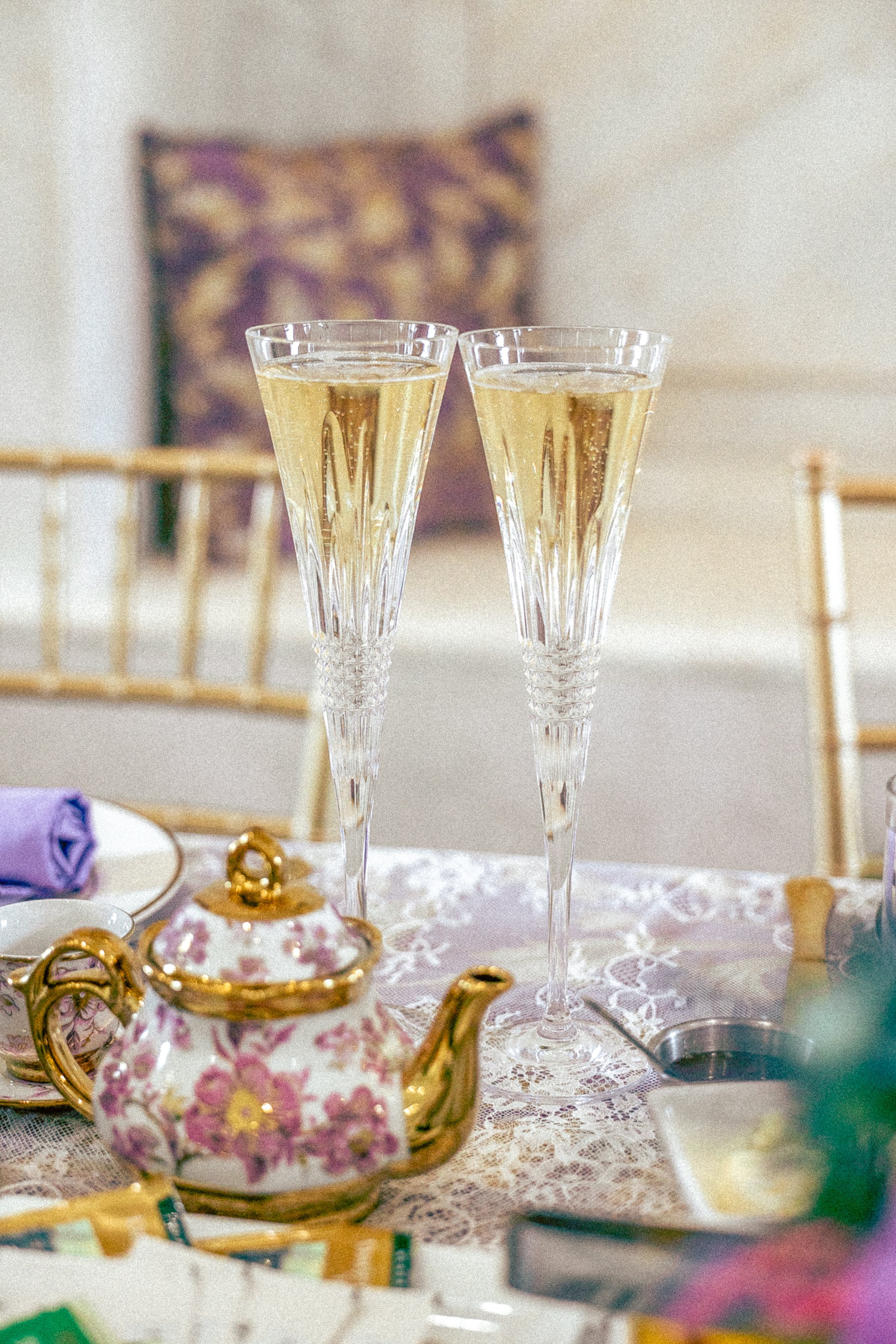 Two champagne flutes filled with sparkling wine or champagne on a lace tablecloth, with a blurred background and decorative items on the table.
