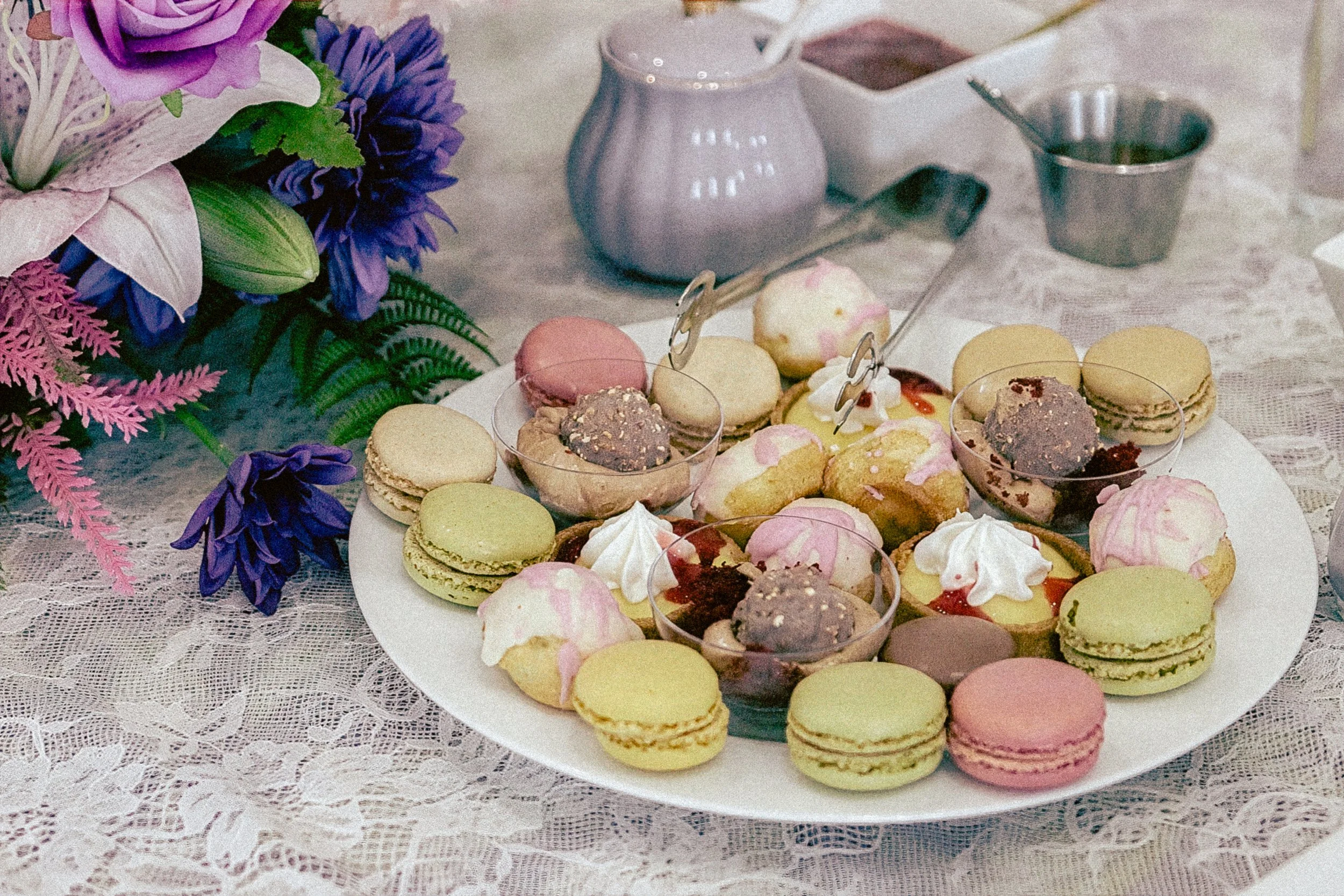 A large white plate filled with colorful macarons, cupcakes, and small desserts, placed on a lace tablecloth. In the background, there are pink and purple flowers, and various pitchers and containers for drinks.