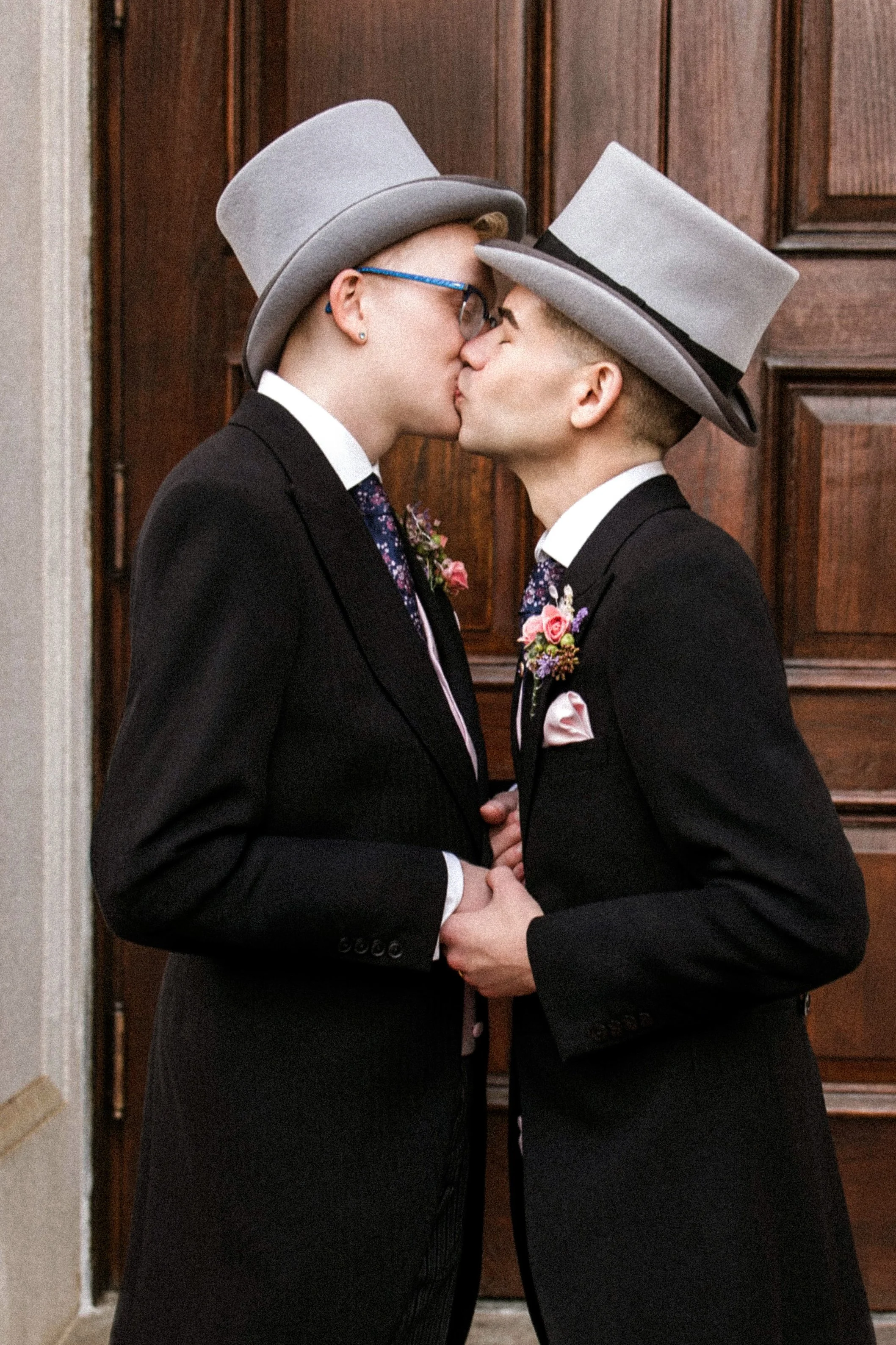 Two people in formal suits and gray top hats kissing each other, holding hands, with a dark wooden door in the background.