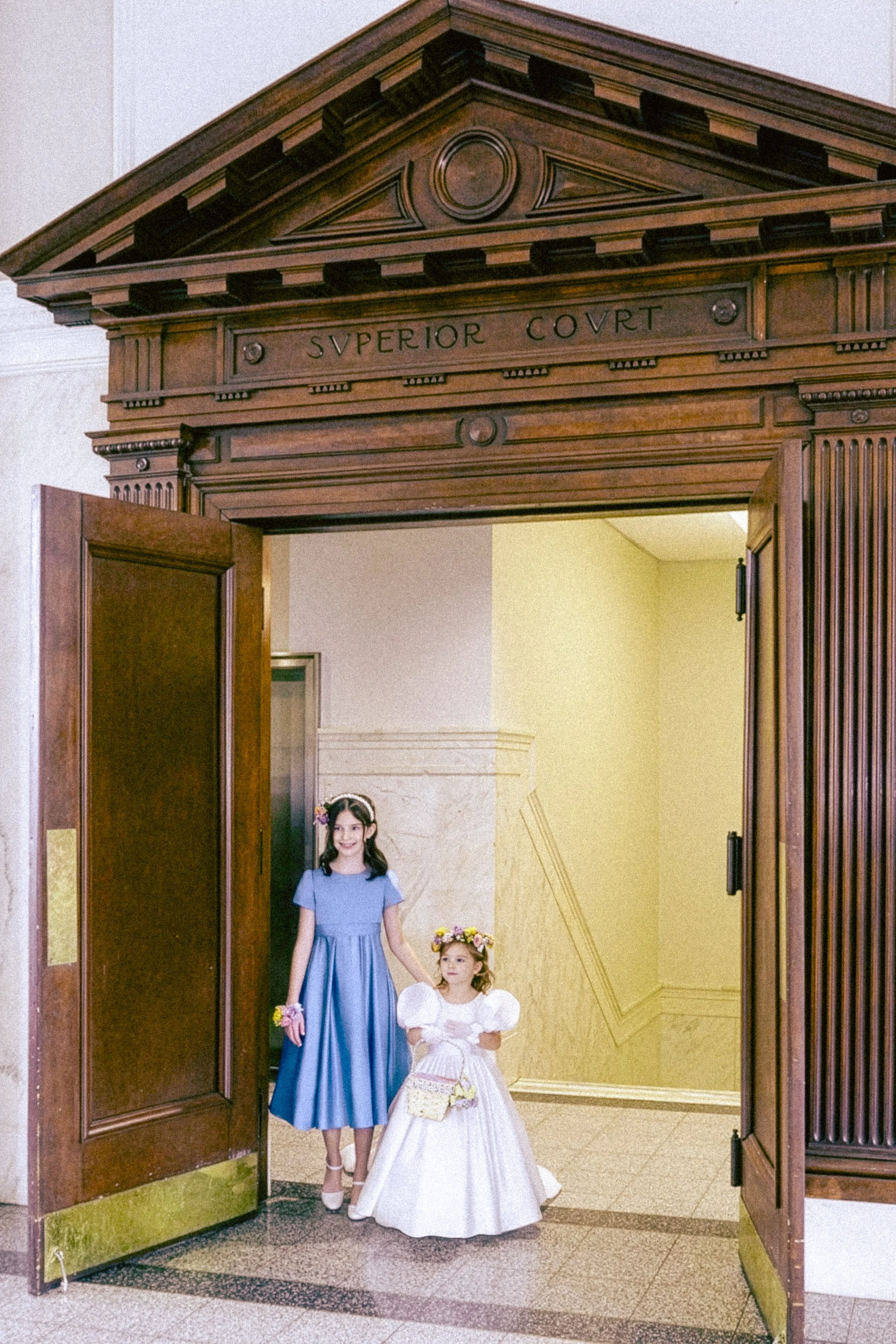 Two young girls in formal dresses standing in a doorway, one in a blue dress holding a dress-up girl, the other in a white dress with floral headband, inside a building with a decorative wooden arch and a yellow wall.