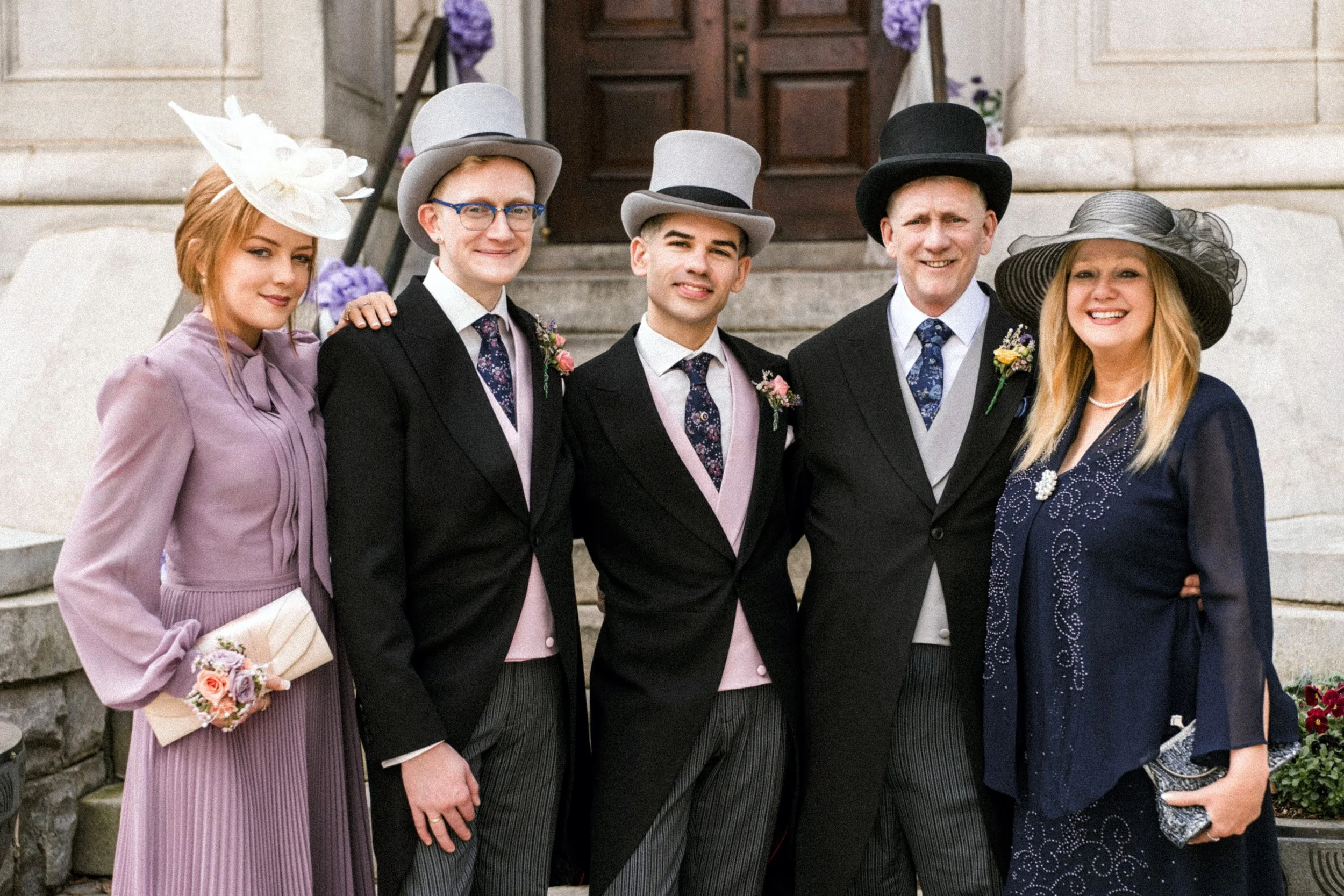 Group of six people dressed in vintage formal attire, standing on the steps in front of a building with wooden door, posing for a photo.