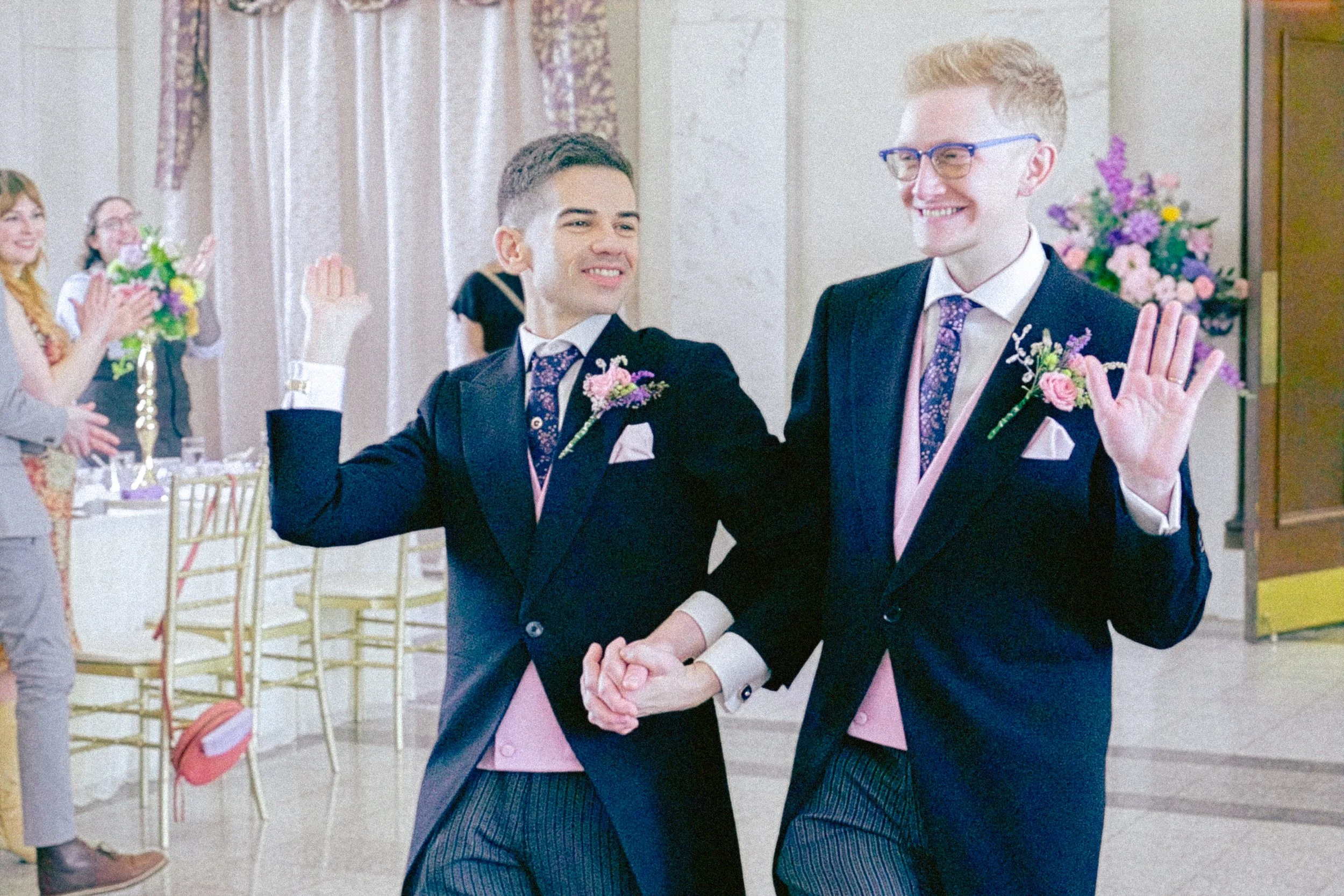 Two men in formal attire, exchanging wedding vows at a wedding ceremony in an elegant venue with floral decorations, smiling and holding hands together.