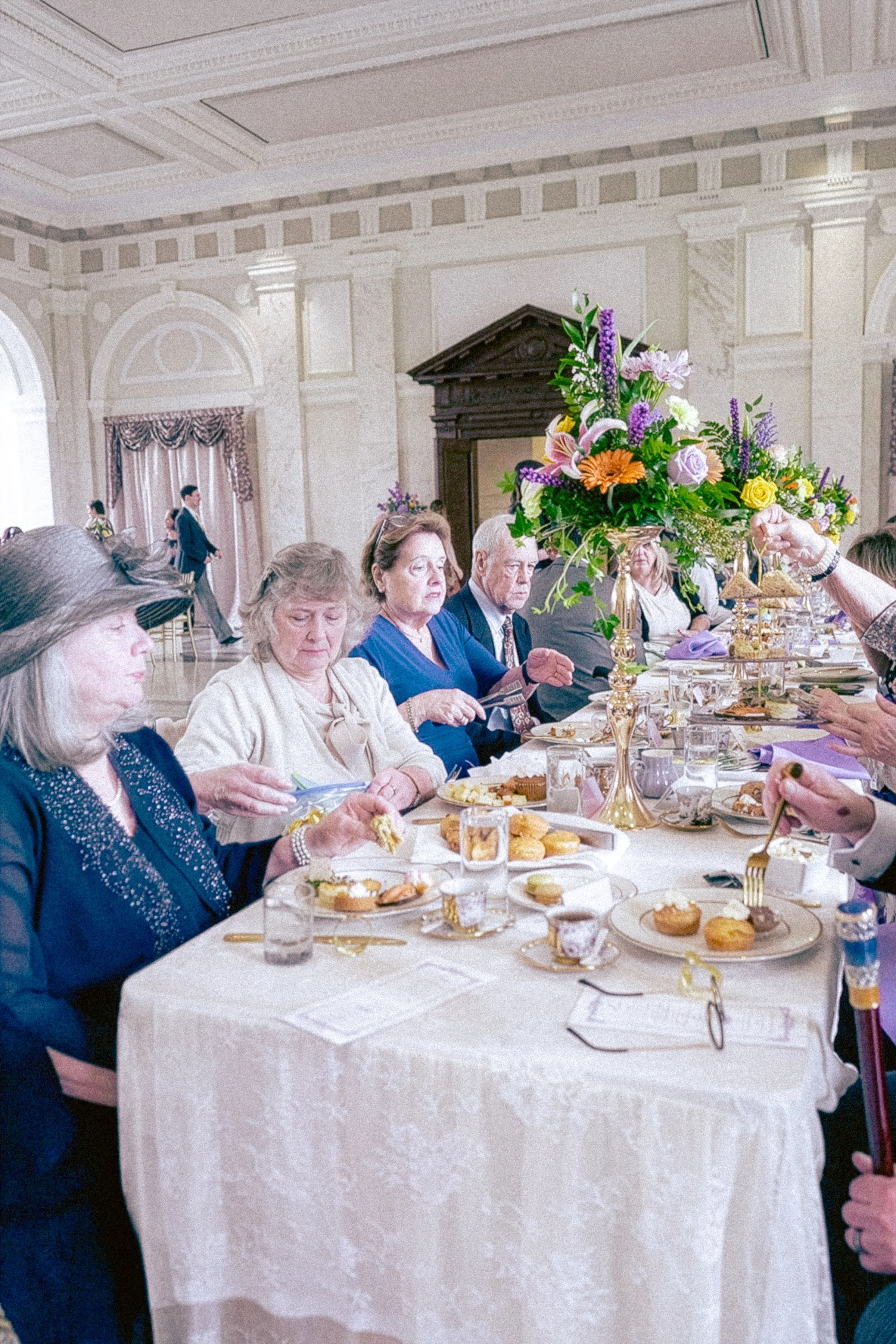 People sitting at a long dining table with plates of food, a large floral centerpiece, and elegant decor in the background.