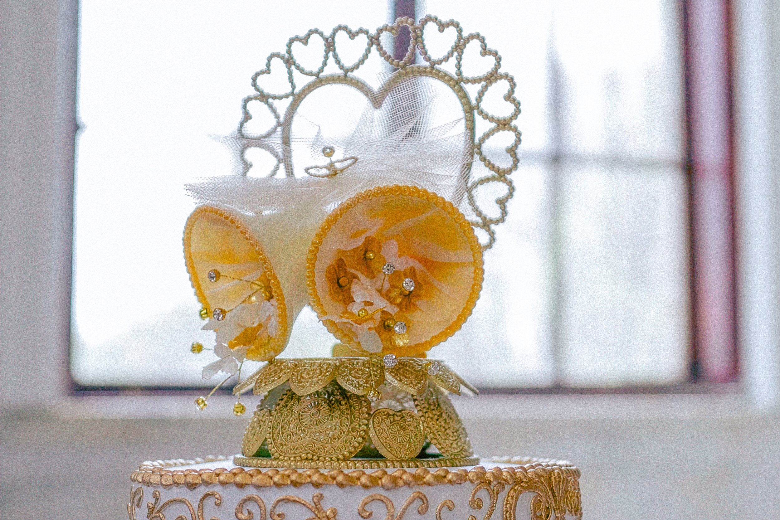Decorative item made of gold, white, and pink materials displayed on a stand with intricate designs, adorned with small gemstones, set against a bright background.