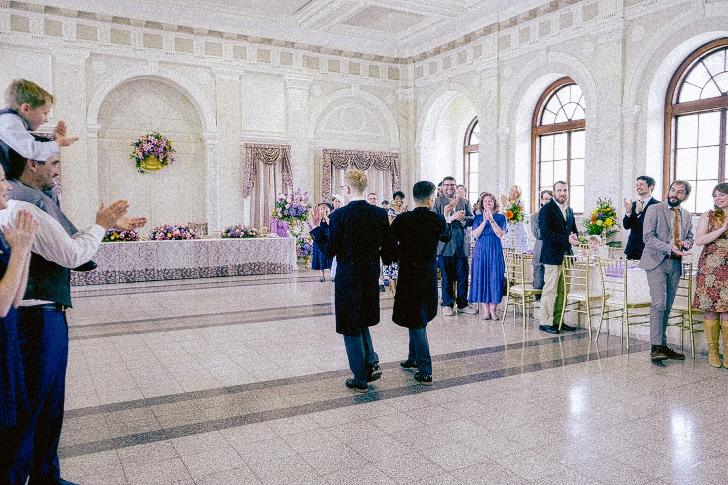 People attending a wedding reception in a grand hall with large arched windows and high ceilings, some clapping and smiling.
