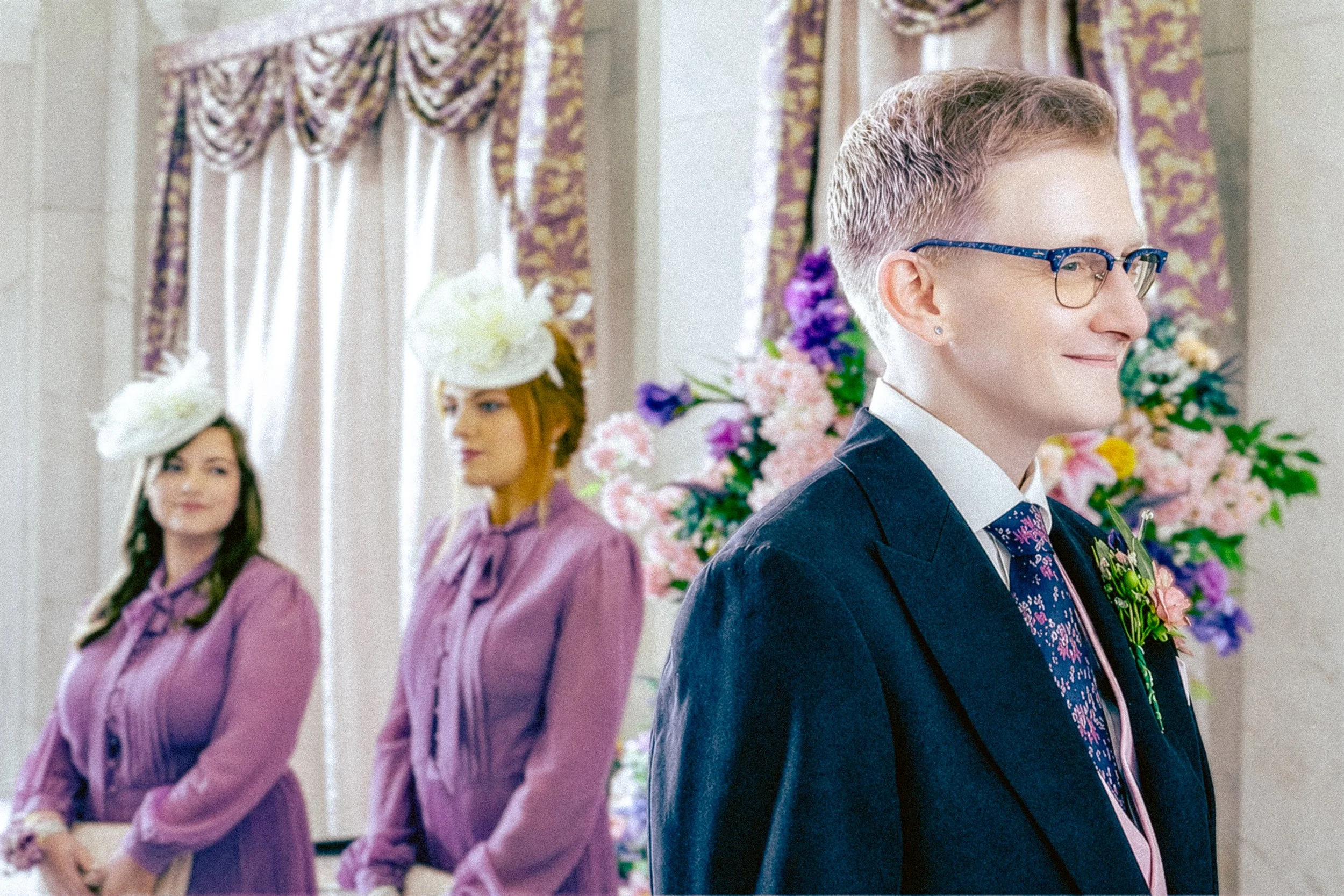 Three women and one man are standing in line, dressed elegantly with floral accessories for a formal event in a decorated room with floral curtains and arrangements.