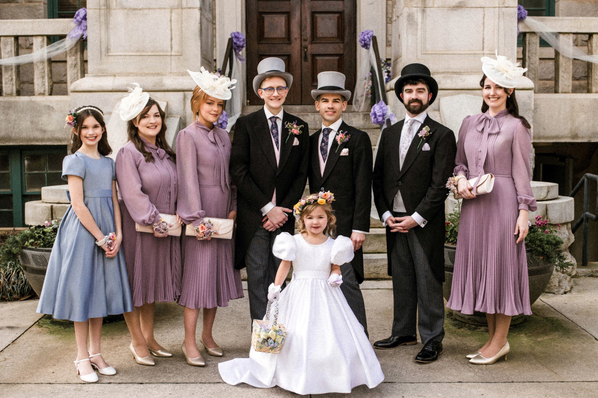 A wedding party standing outside a stone building with wooden doors. The group includes young girls in pastel dresses and women in matching purple dresses with large hats, along with two men and a woman in dark suits and hats. The bride is a young girl in a white dress holding a basket, with the rest of the group smiling and posing for the photo.