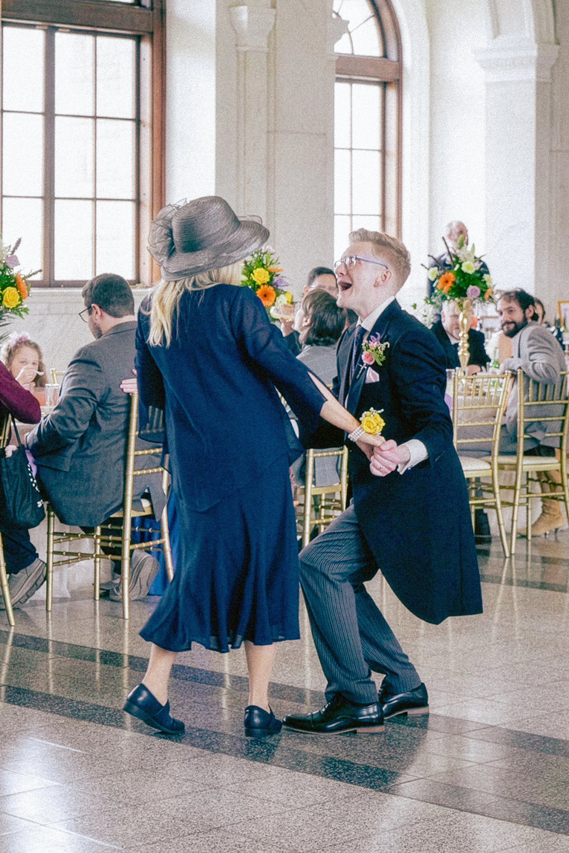 A couple dancing together at a wedding reception in a large room with tall windows and floral decorations on tables. The woman is wearing a navy dress and a wide-brimmed hat, and the man is wearing a dark suit with glasses, smiling and holding hands.