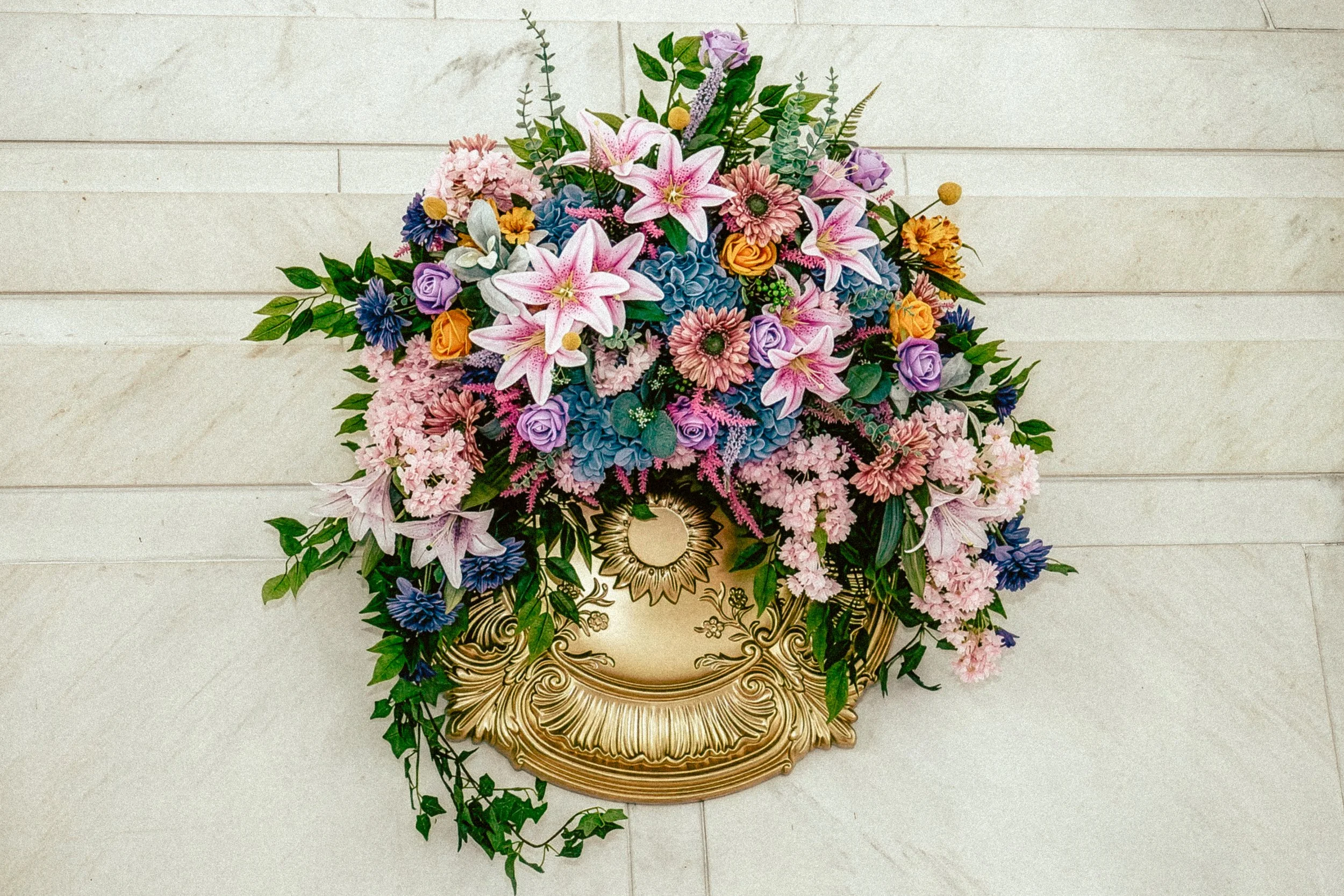 A large floral arrangement with pink lilies, purple roses, blue hydrangeas, and various greenery in an ornate gold and black vase on a light-colored tiled floor.