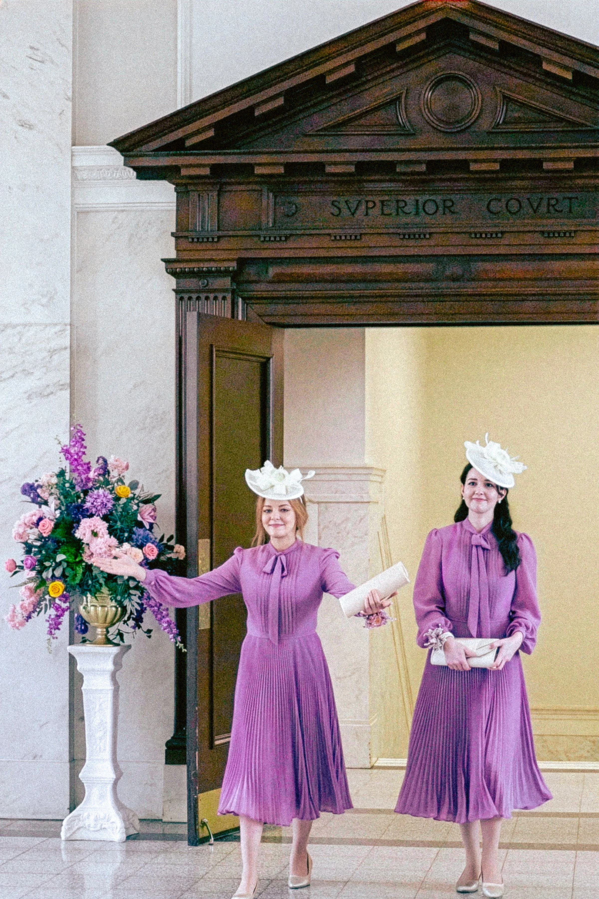 Two women dressed in purple pleated dresses with matching hats, standing indoors near a floral arrangement on a white pedestal. One woman holds a bouquet of colorful flowers and a rolled-up paper, while the other holds a book or folder. They are wear
