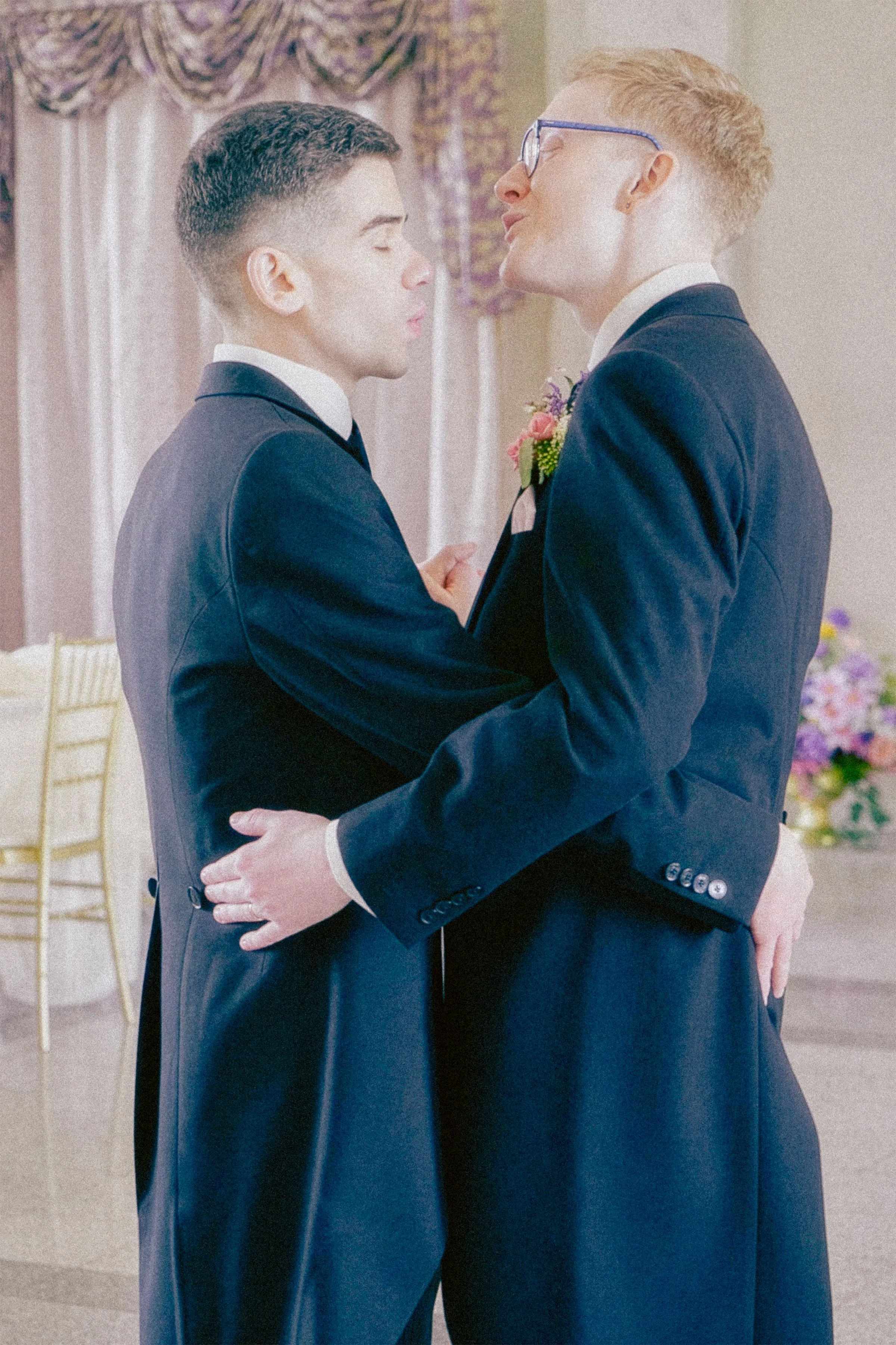 Two men in tuxedos are dancing closely at a wedding reception, with a decorative curtain and floral arrangement in the background.