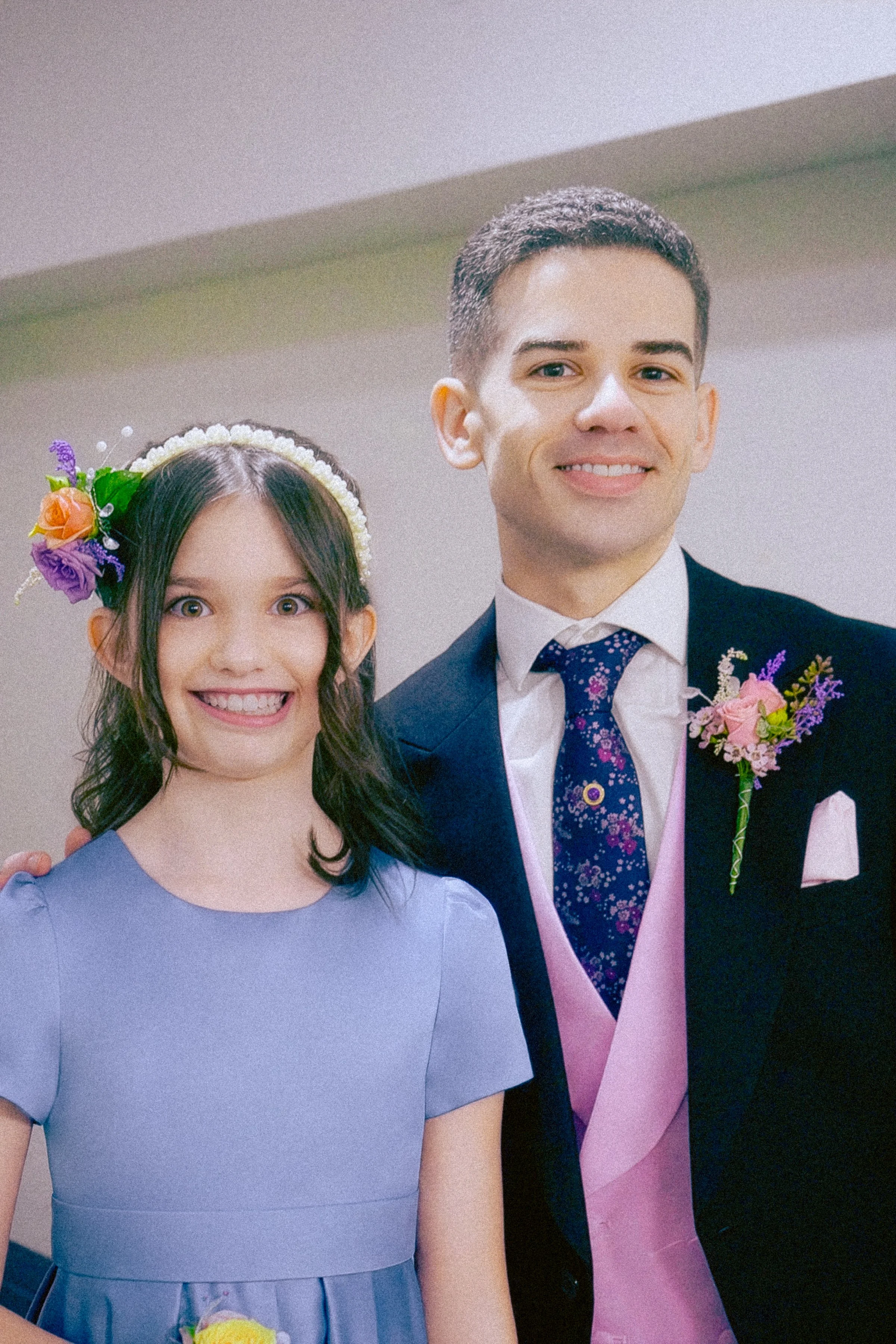 A young woman and a young man dressed formally, smiling for a photo at a special event. The woman is wearing a lavender dress and a floral headband, and the man is in a dark suit with a patterned tie and a pink sash, with a boutonniere.