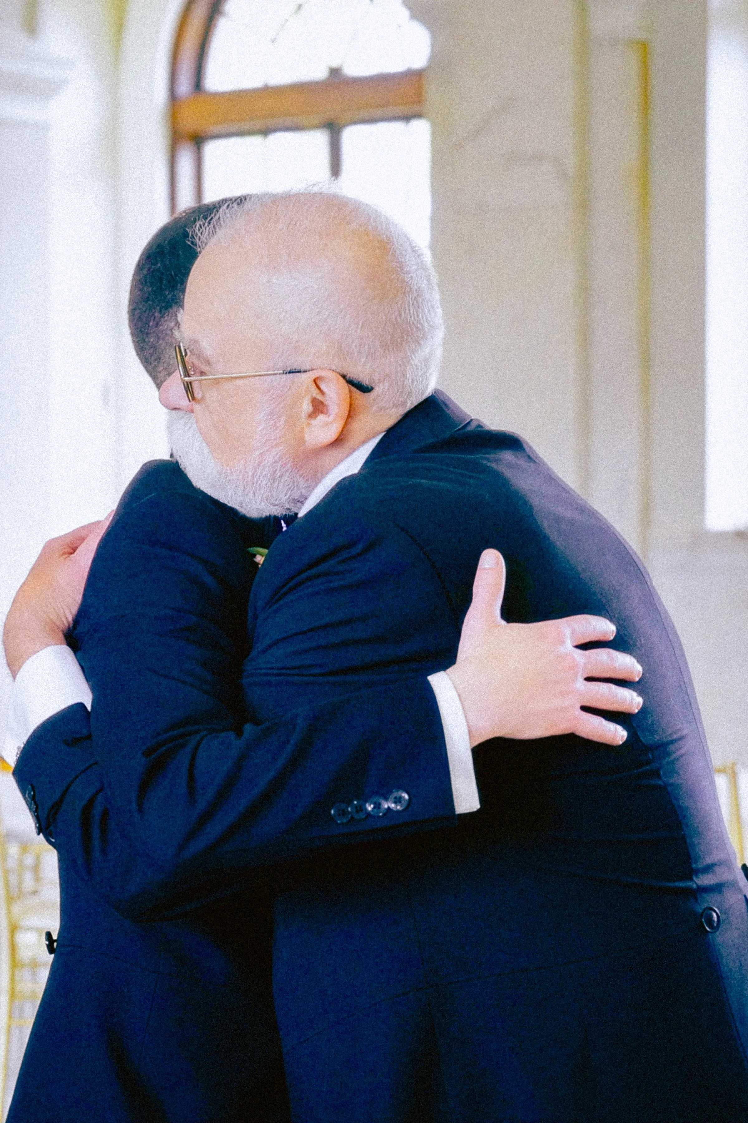Two men in suits hugging each other warmly indoors near a window.