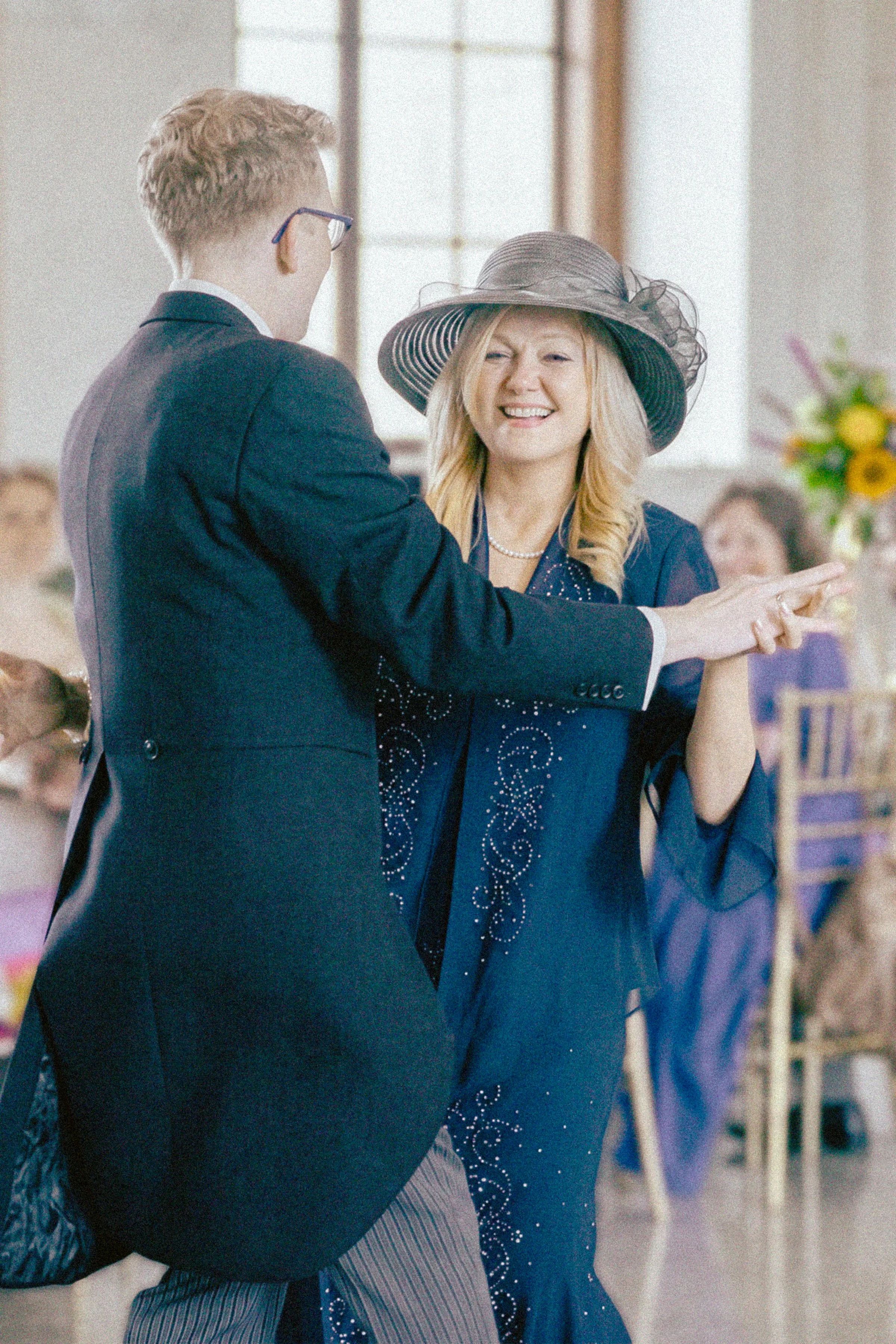 A young man in a navy suit and glasses is dancing with an older woman in a blue dress and large floppy hat at a social event. She is smiling and they are holding hands.