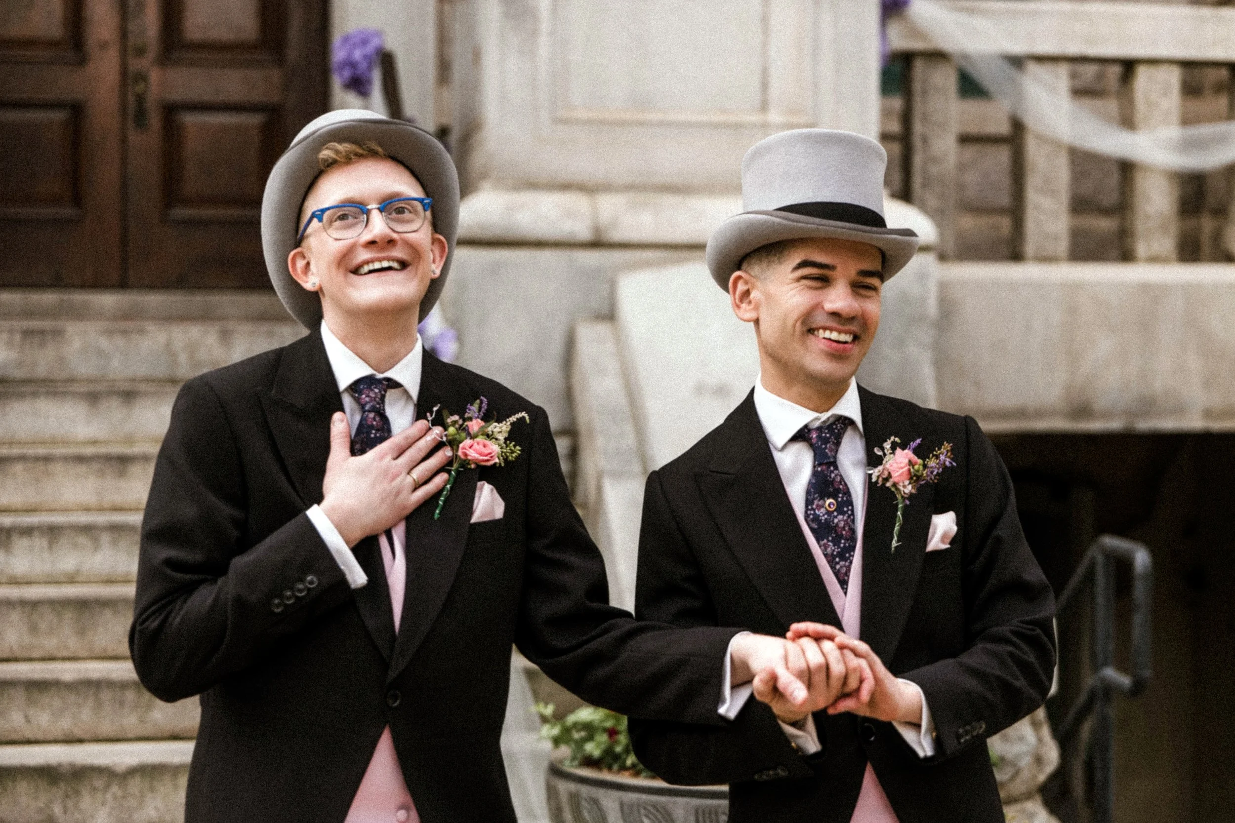 Two men dressed in tuxedos and pink vests, standing outside, wearing gray top hats with black bands, during a wedding ceremony. The man on the left is smiling and has his hand on his chest, while the man on the right is smiling with their hands clasp