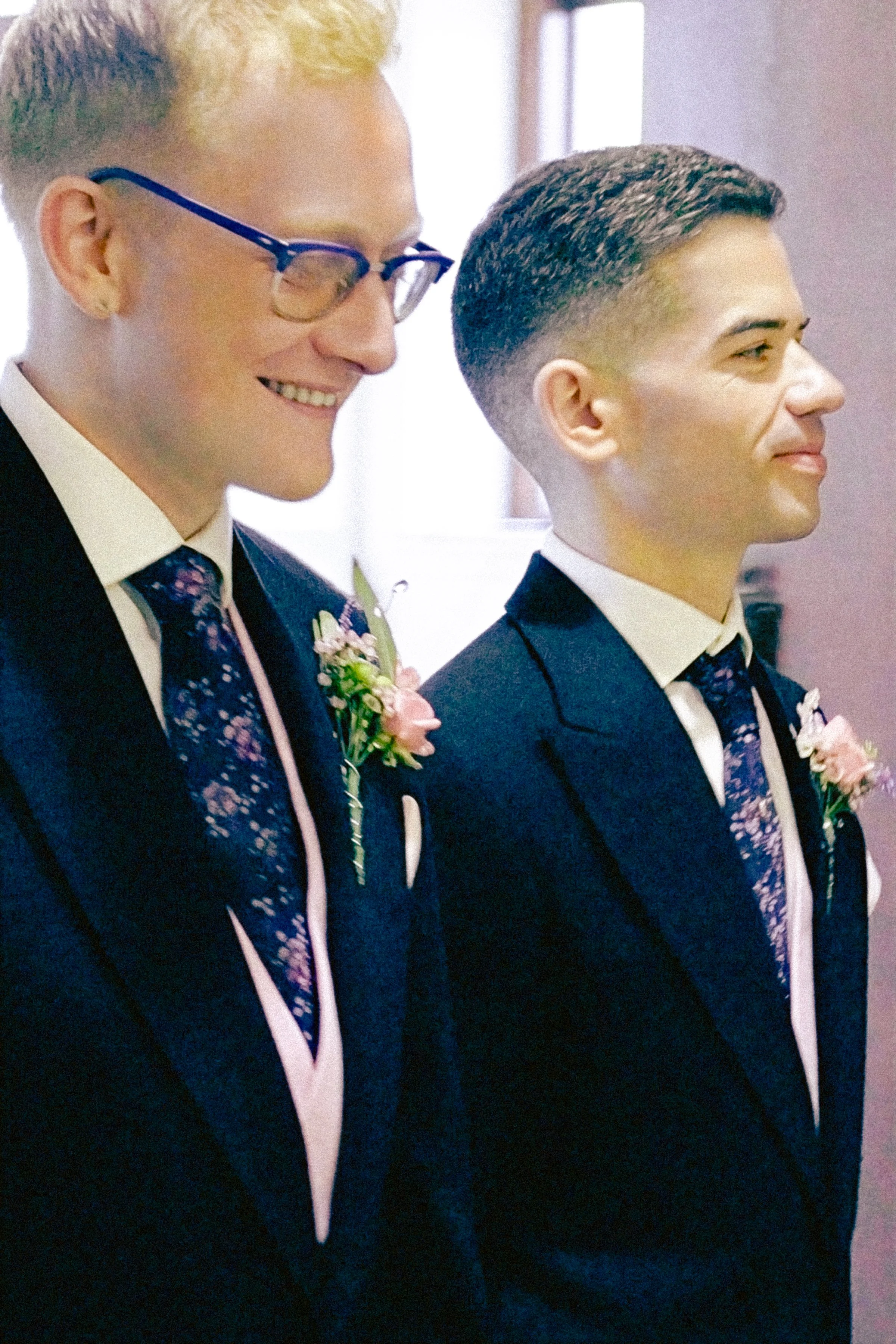 Two young men dressed in formal suits, smiling and standing side by side in a ceremony setting.
