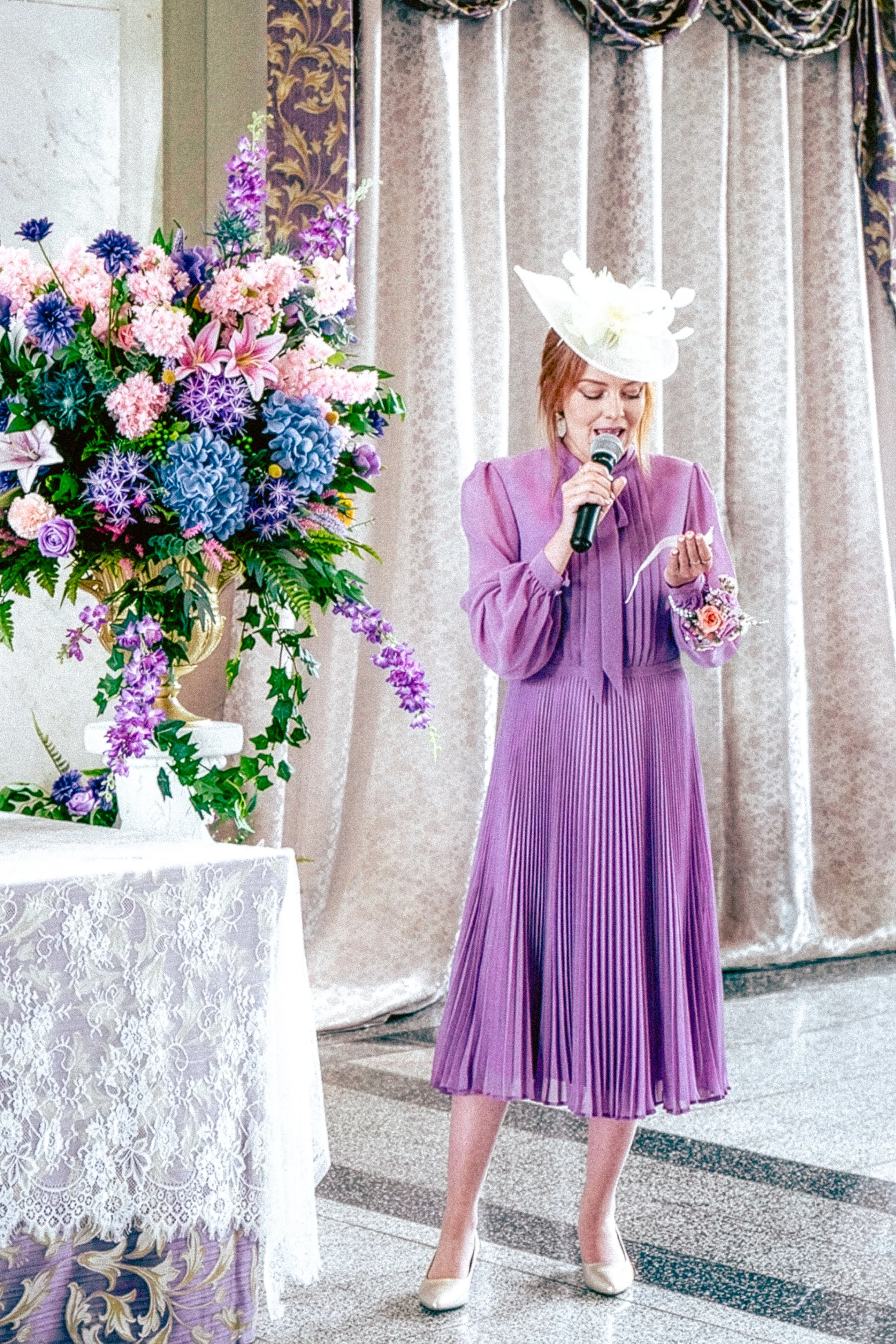 A woman dressed in a purple pleated dress and white shoes is speaking into a microphone at a formal event. She's wearing a white hat with a large flower and has a corsage on her wrist. To her left, there's a large floral arrangement of pink, purple, 