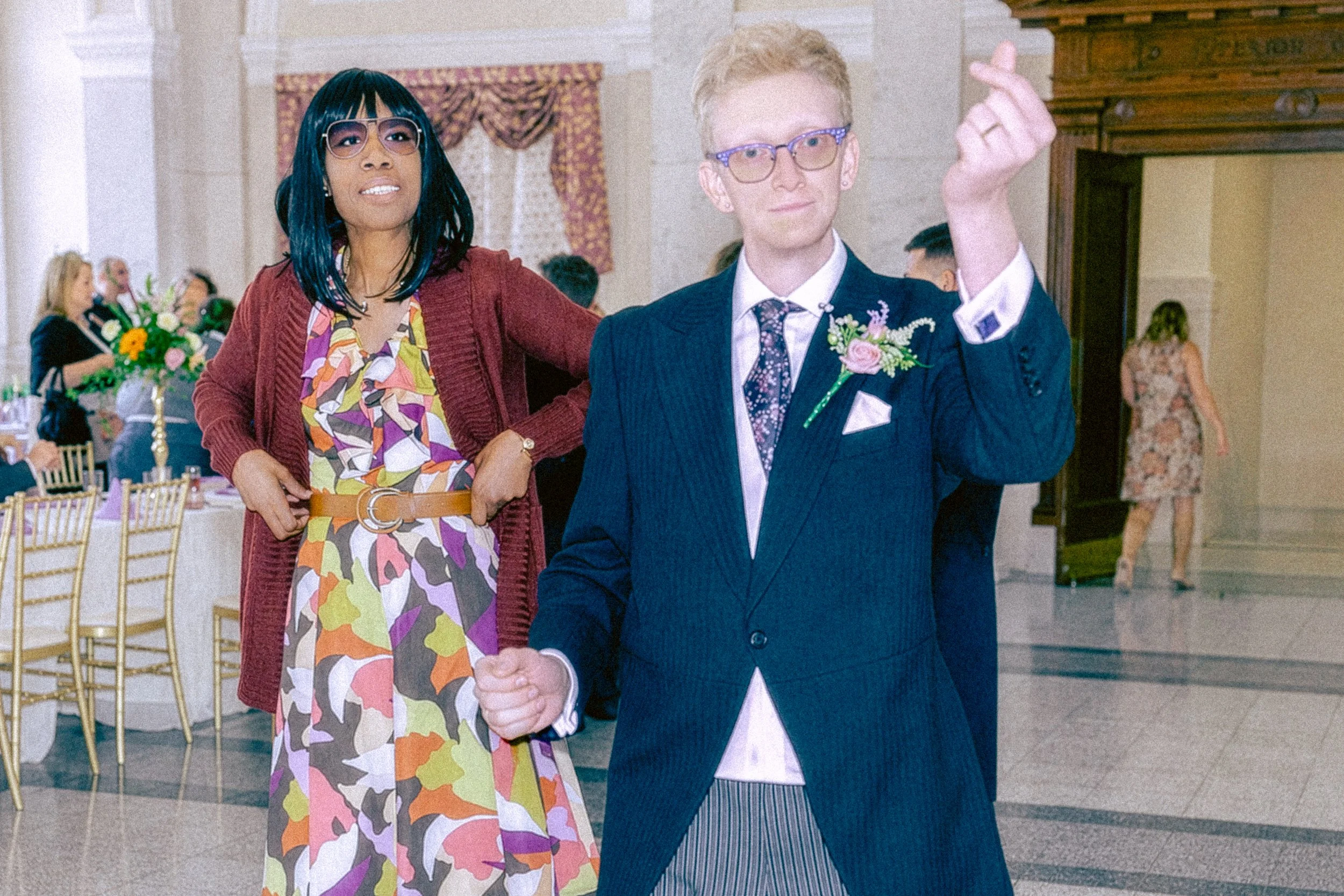 A woman and a man shaking hands at a wedding reception. The woman has glasses and dark hair with teal streaks, wearing a colorful patterned dress and a red cardigan. The man with blonde hair and glasses is dressed in a navy suit with a floral boutonn
