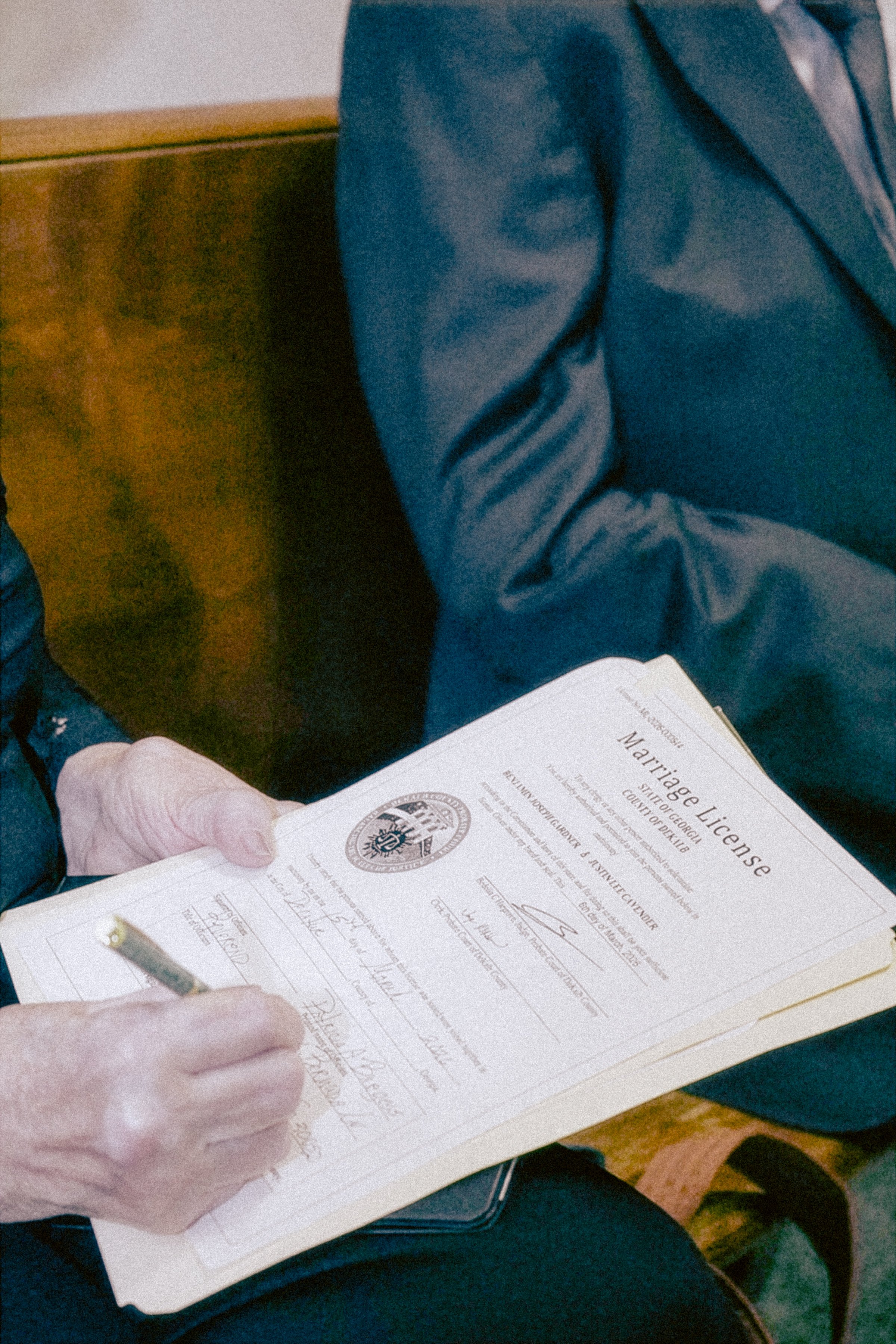 Person in a suit holding a marriage license and signing it, with a pen in hand, sitting at a wooden table.