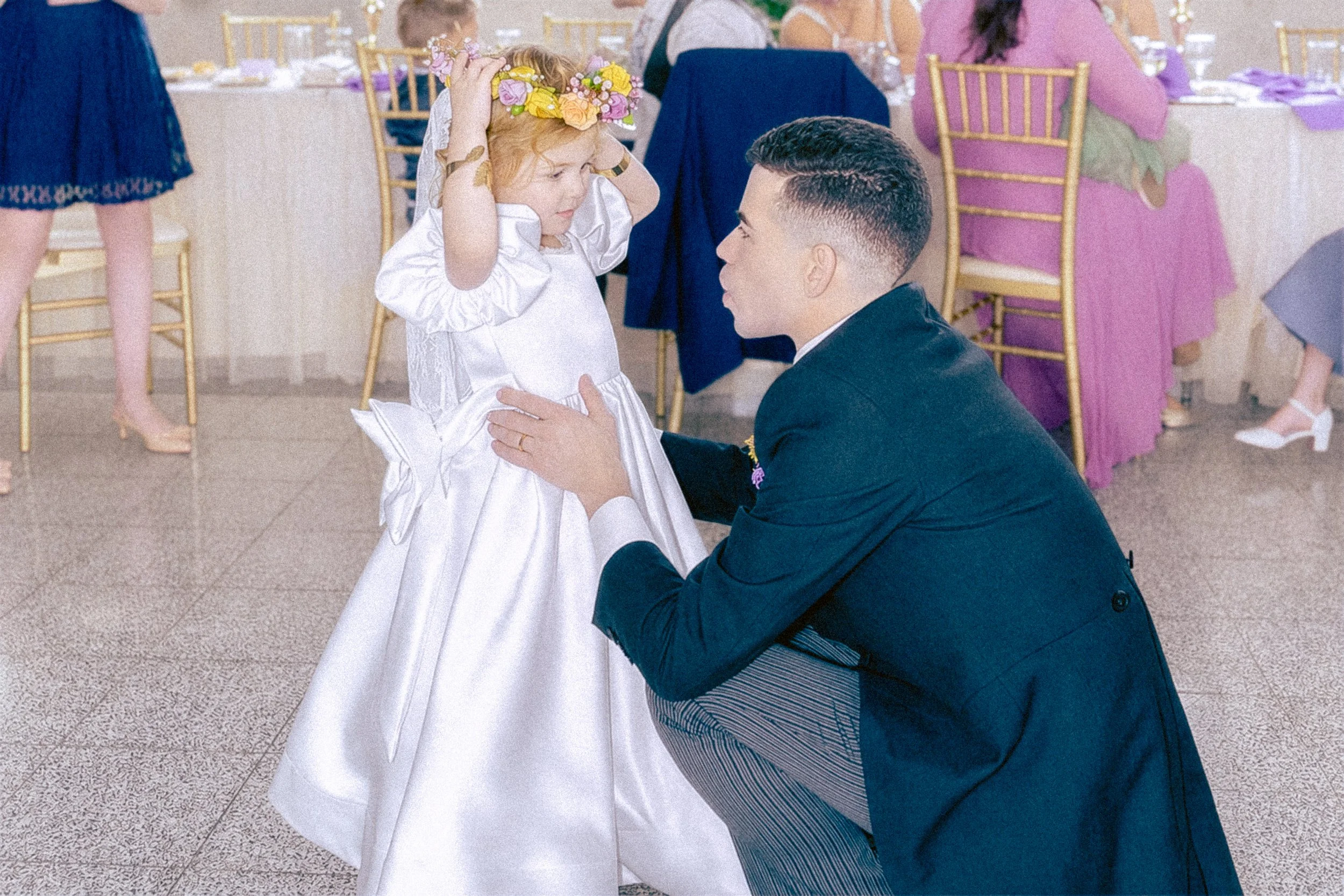 A young girl in a white dress with a flower crown on her head is being kneeling by a man in a suit at a celebration or wedding reception. The girl is adjusting her flower crown while the man looks up at her, smiling. The background shows decorated ta