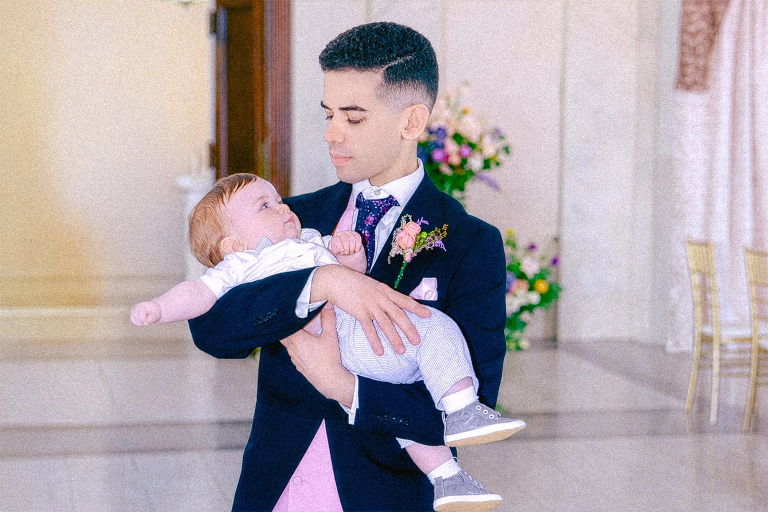 A man in a suit holding a young boy, both smiling at each other, indoors with curtains and a floral arrangement in the background.