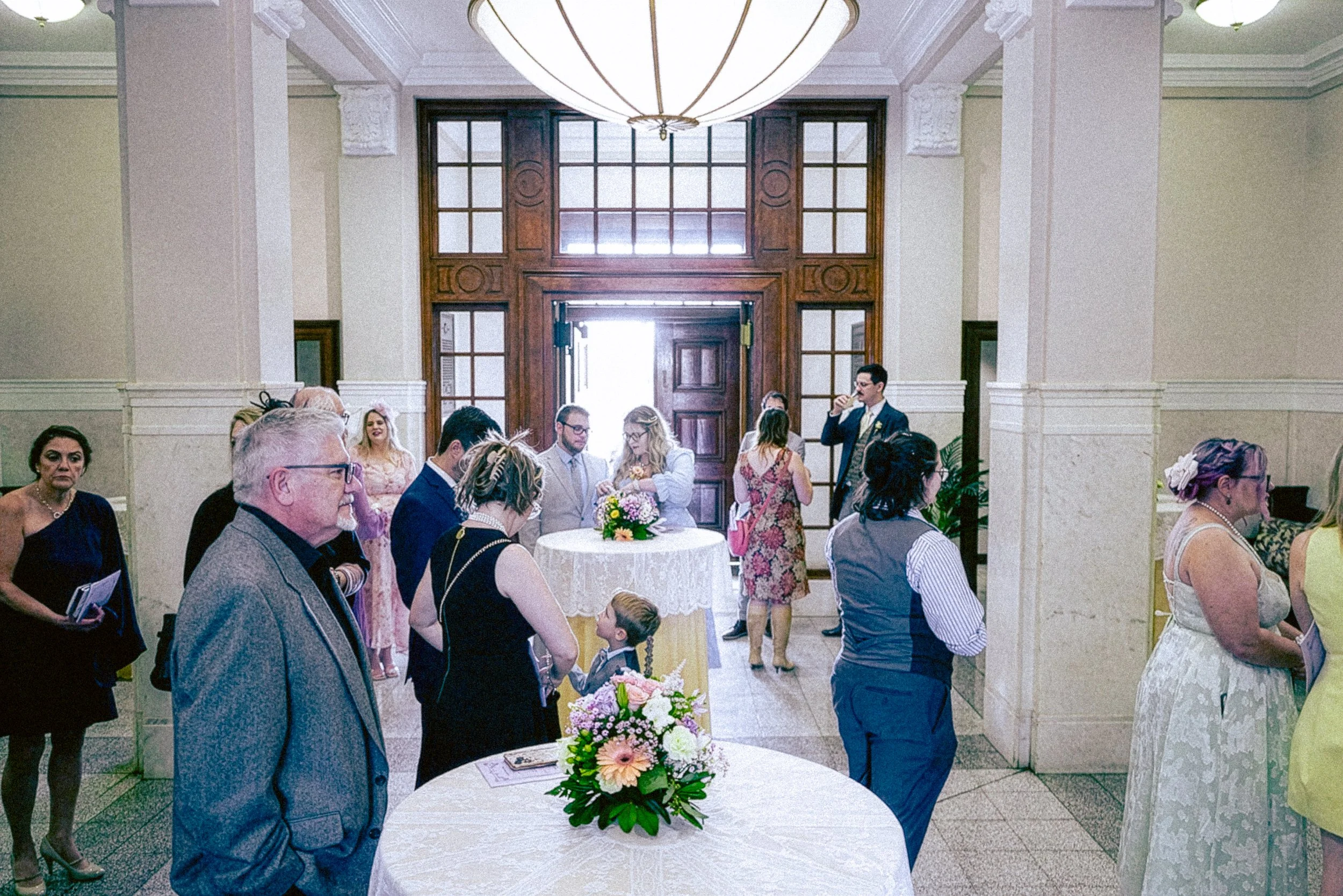 People gathered around a table with a bouquet of flowers in a bright, elegant room with high ceilings and a wooden door in the background at a social event.