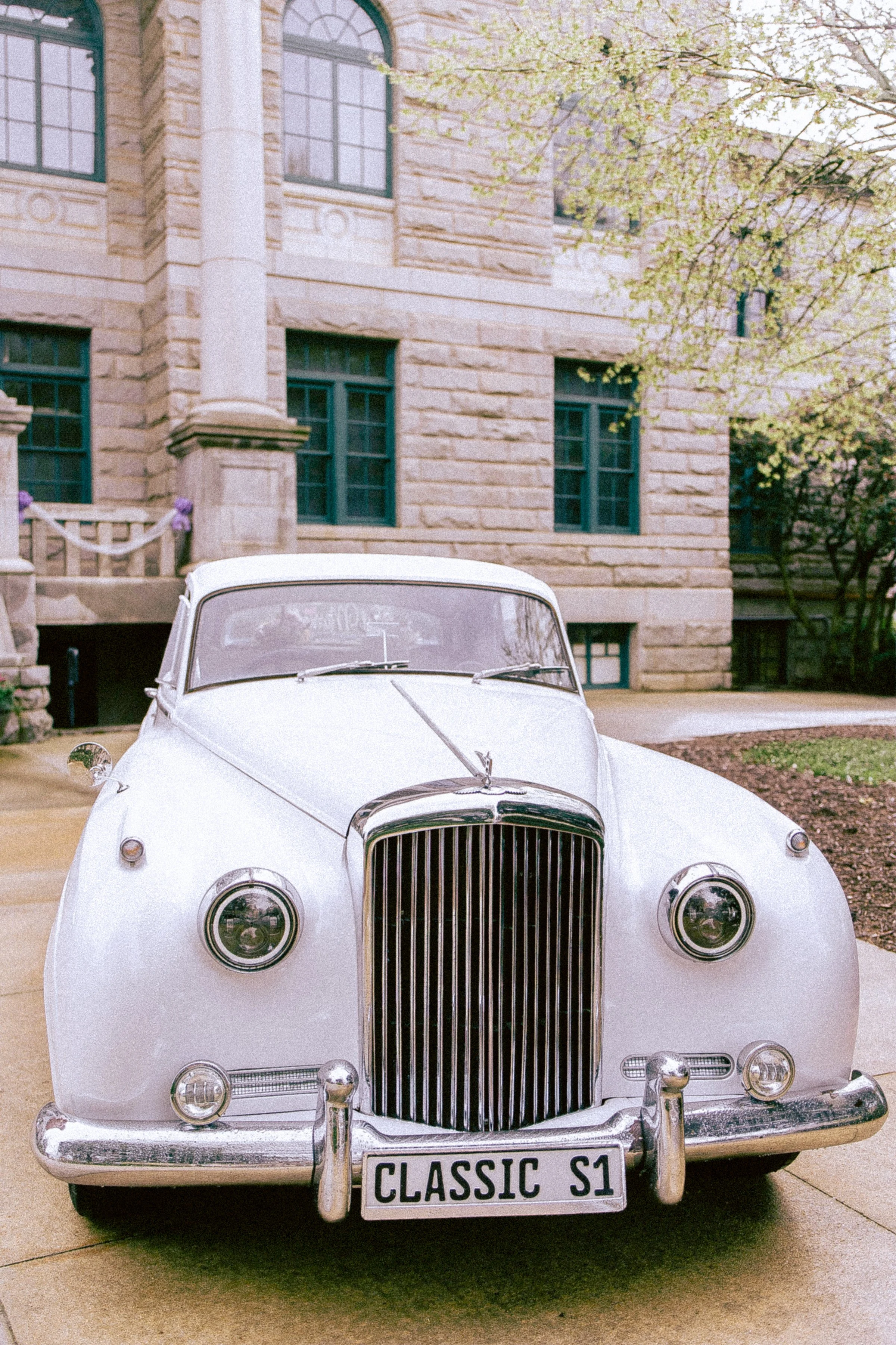 Front view of a vintage white car parked in front of a stone building with multiple windows, surrounded by trees with light green leaves.