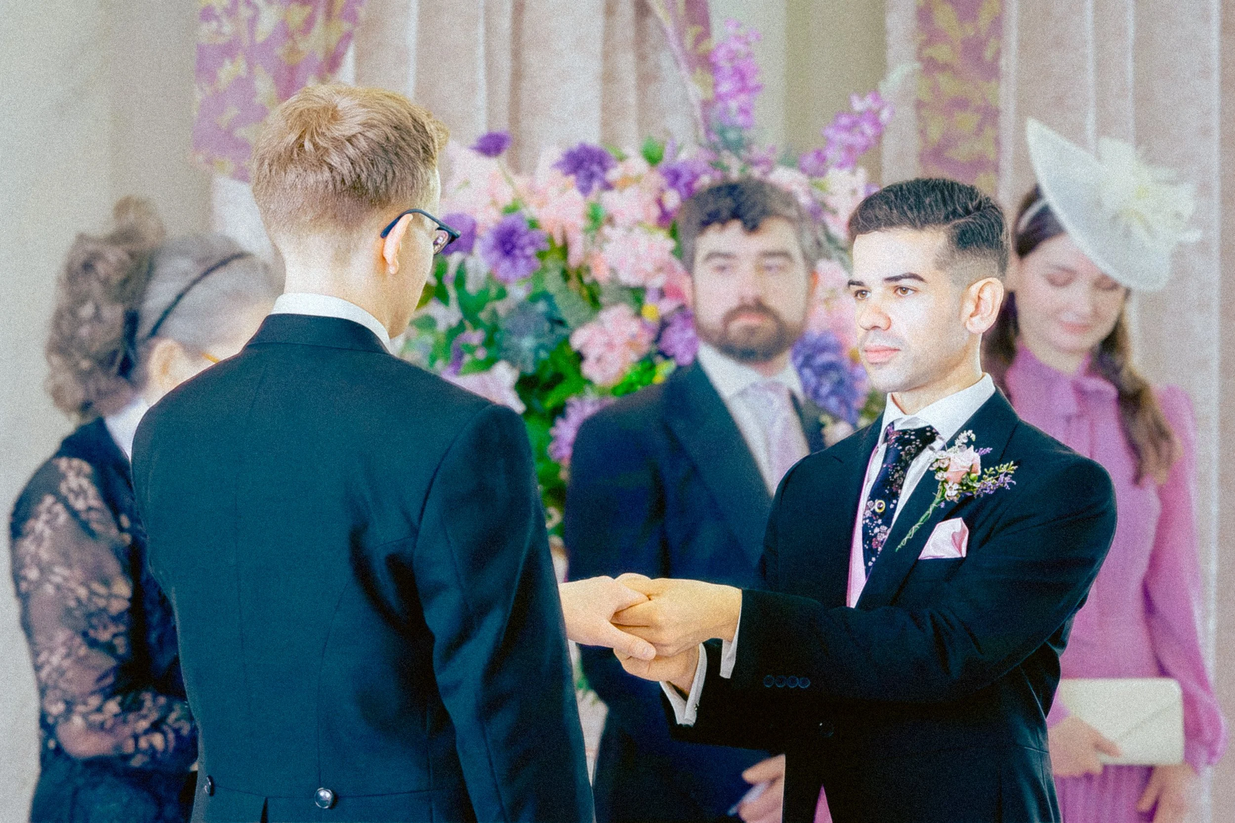 A same-sex couple is exchanging rings during their wedding ceremony, with guests and colorful floral decorations in the background.