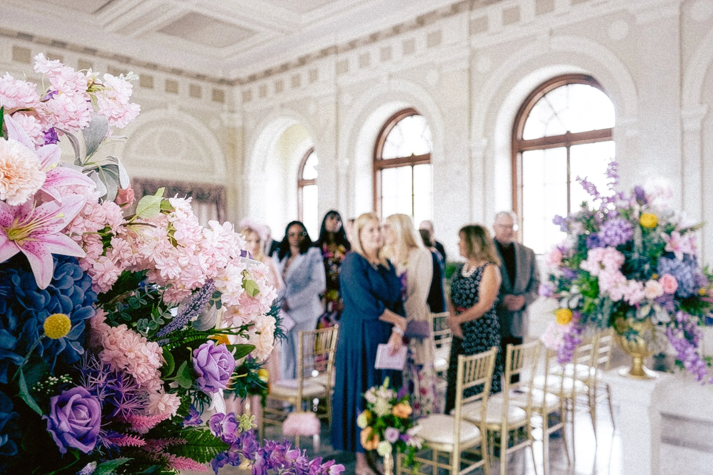 A group of people standing in a room with large arched windows, decorated with colorful flowers, possibly at a wedding or events celebration.