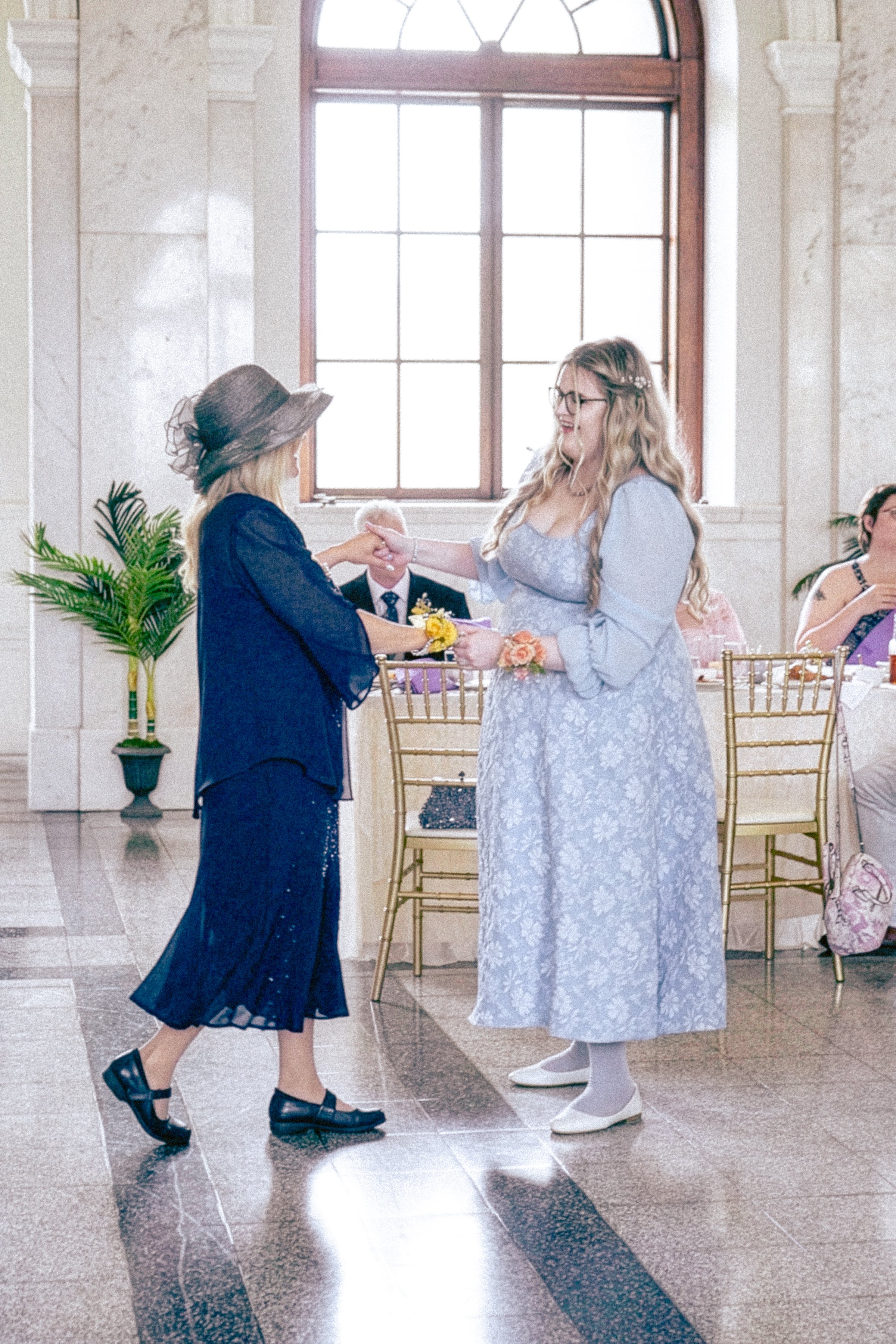 Two women exchanging a handshake at a wedding reception, inside a decorated hall with large window, gold chairs, and potted plants.