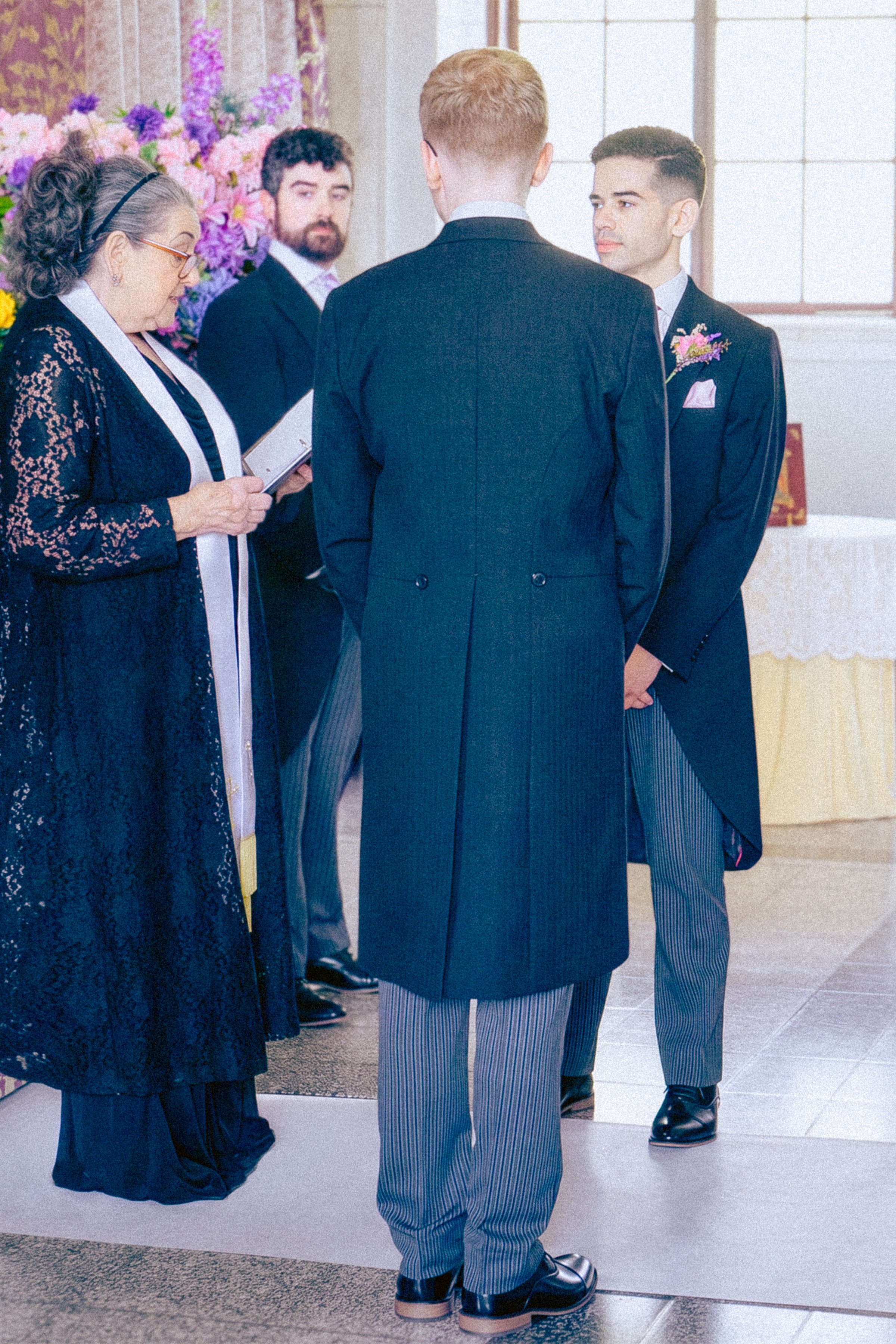 A wedding ceremony with four people, three men and one woman, standing indoors near a floral background. The woman is reading from a booklet, and the men are dressed in suits, with one wearing striped pants and a boutonniere.