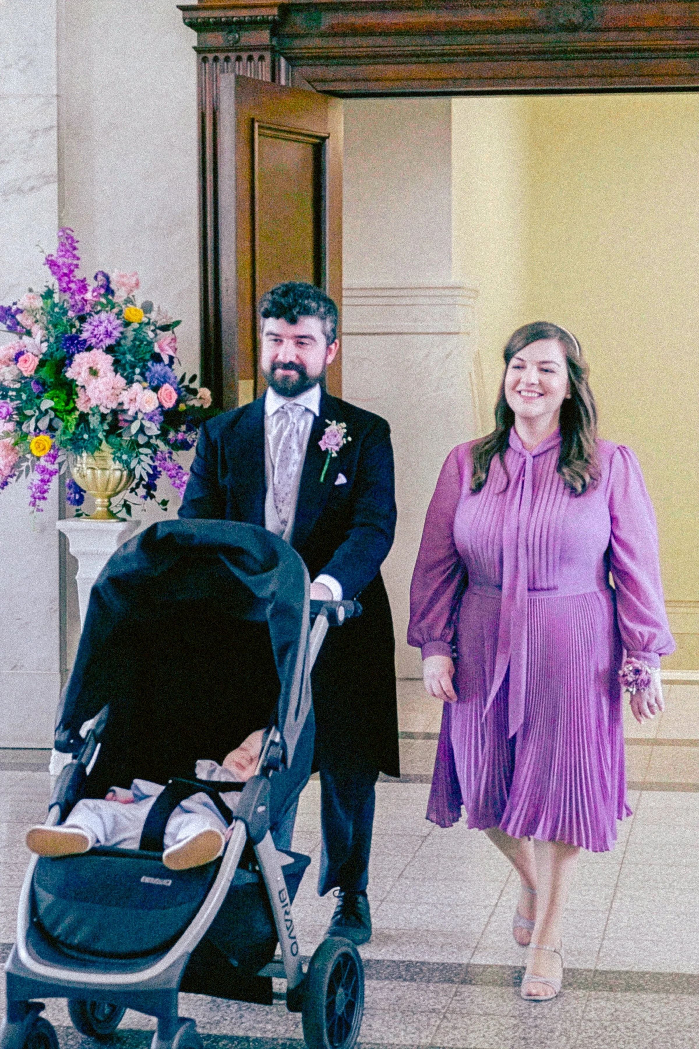 A man with a beard in a formal suit and a woman in a purple dress walking together in an indoor setting, with a baby in a stroller nearby and a large floral arrangement on the side.