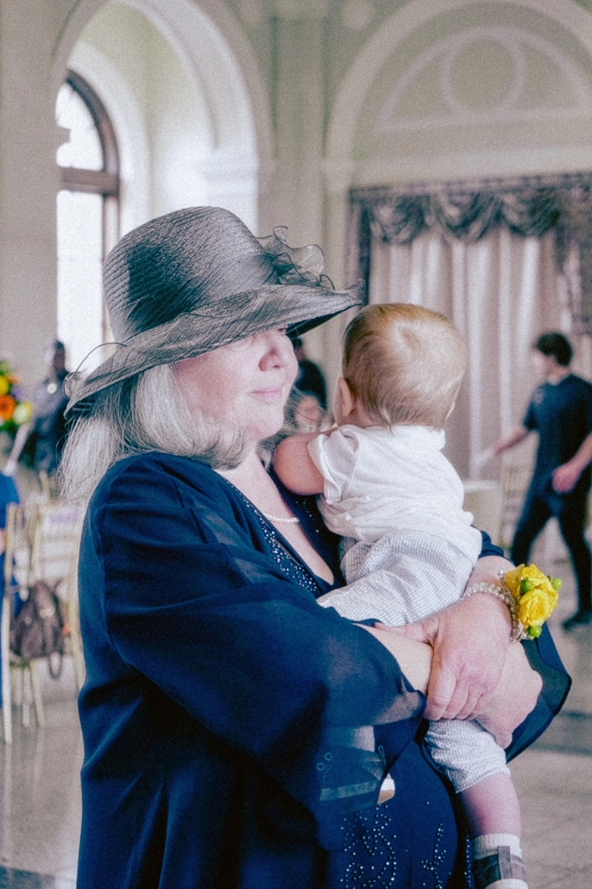 An elderly woman with long gray hair wearing a large gray hat, a navy blue dress with embellishments, and a pearl necklace, holding a young child with red hair dressed in a cream-colored outfit, inside a grand room with high arches, large windows, an