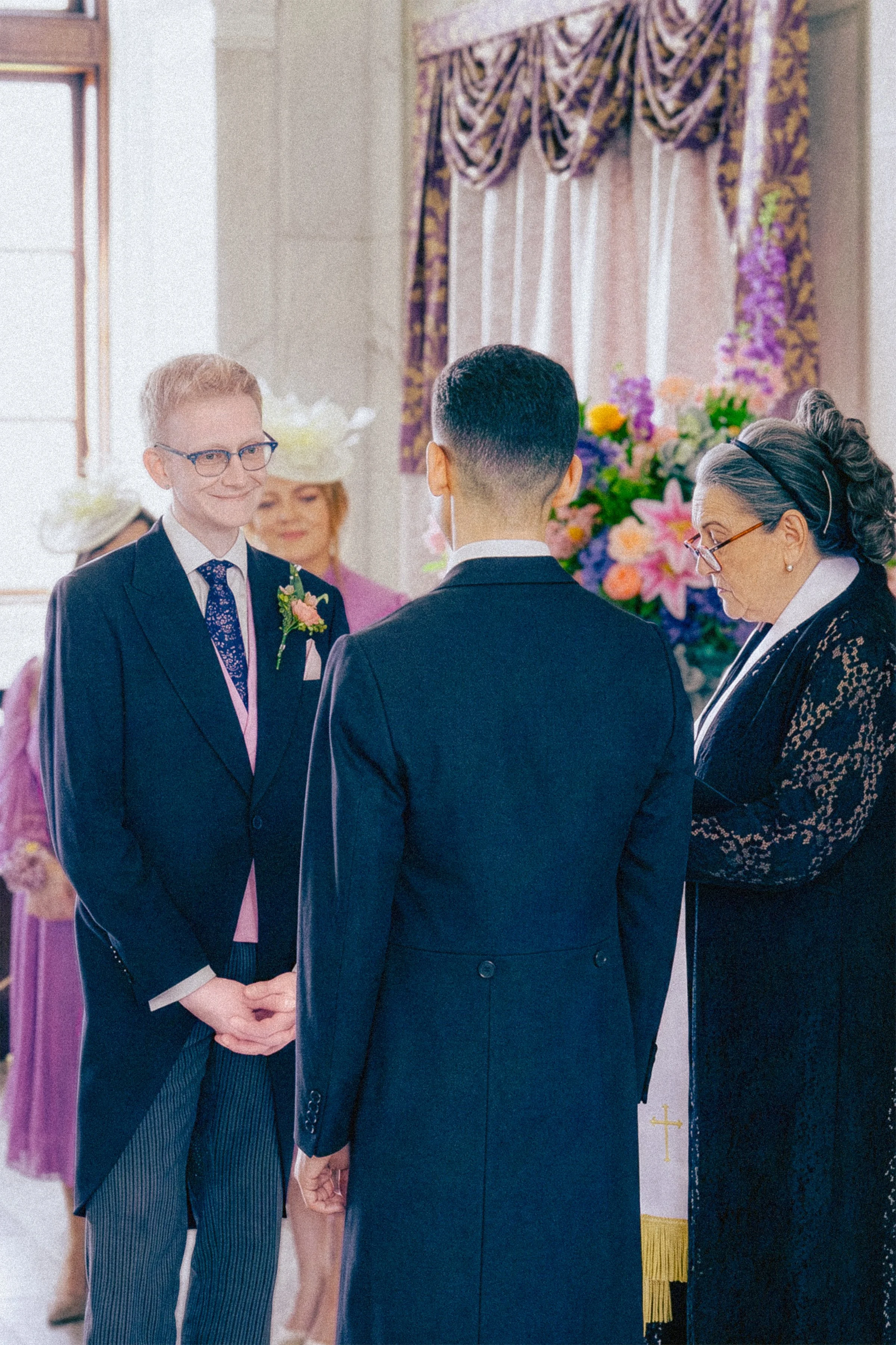 A wedding ceremony with a couple holding hands, facing each other, indoors with floral arrangements and a woman officiant wearing glasses and a black dress with lace details.