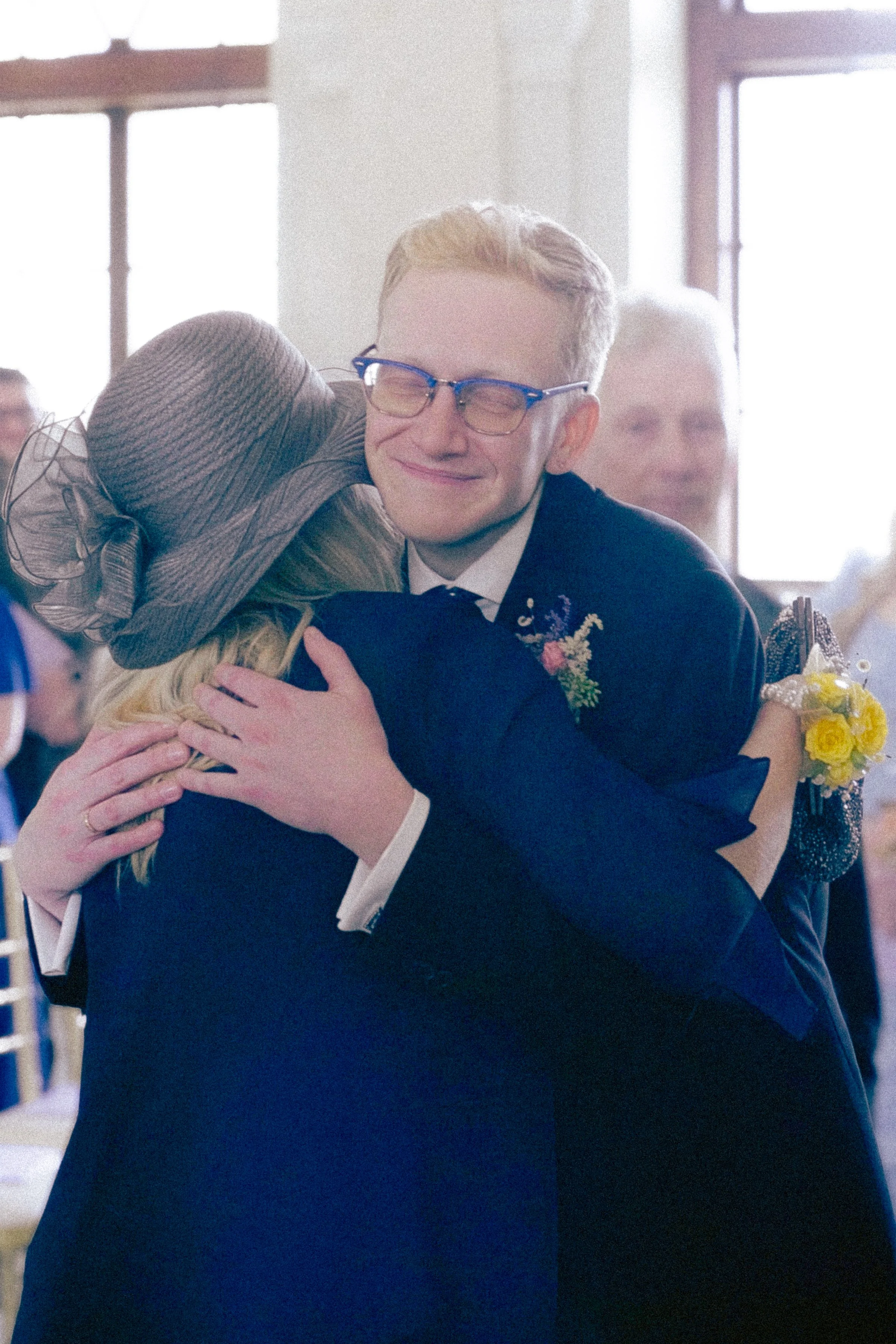 Two people hugging at a formal event, with a man smiling and wearing glasses, and a woman with a wide-brimmed hat.