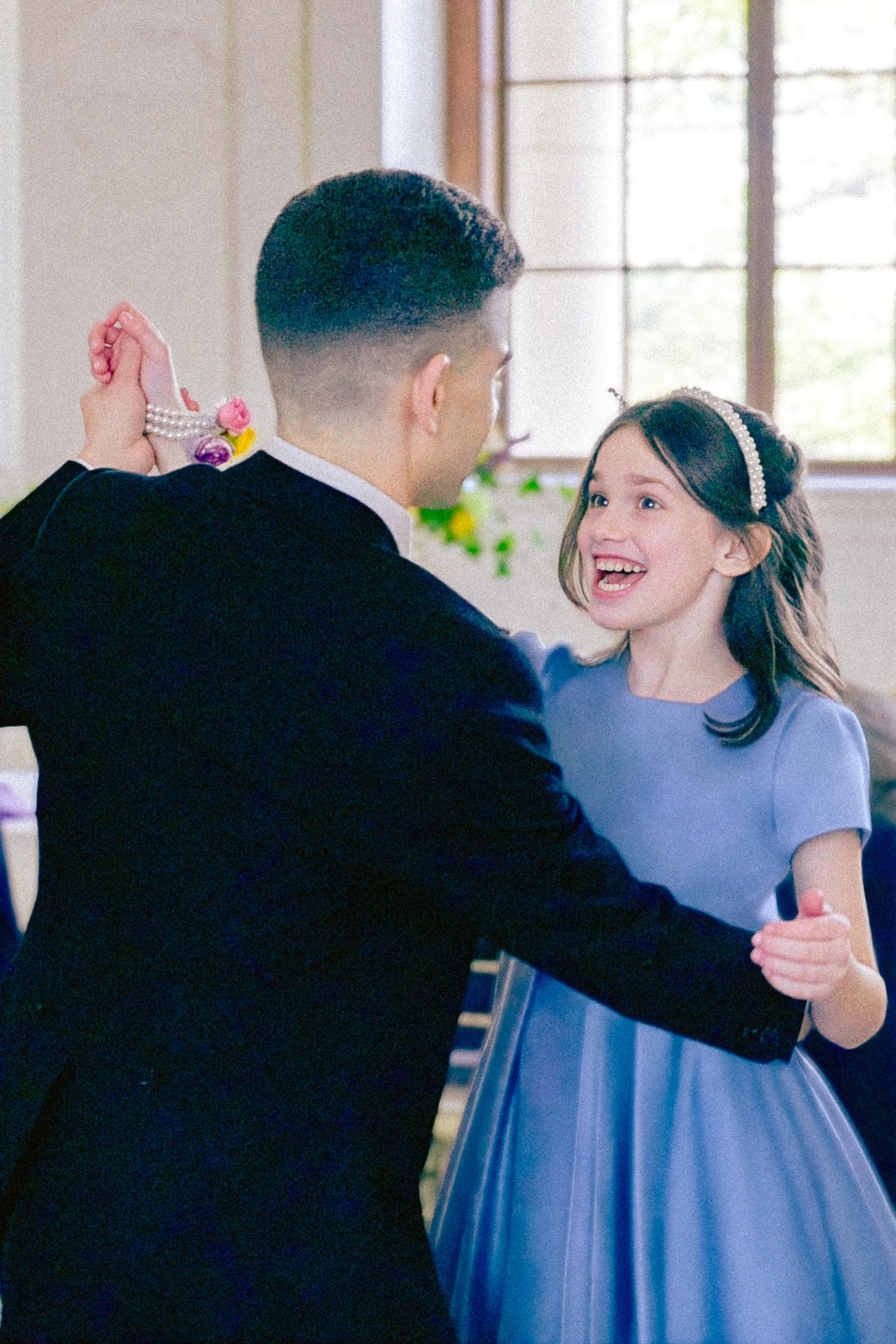 A young girl with a joyful expression dancing with a man in a formal suit, possibly at a dance or special event, with large windows and natural light in the background.