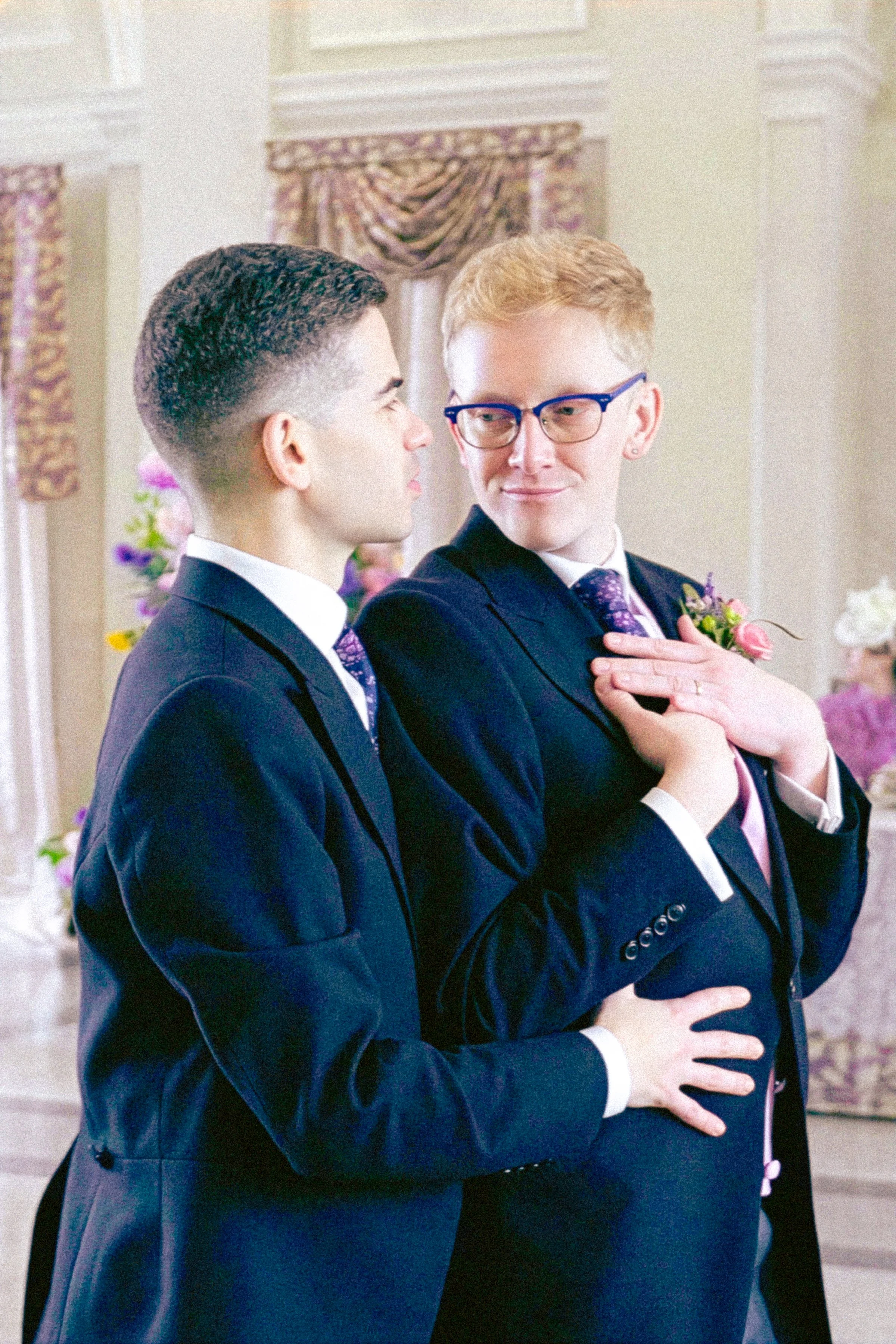 Two men dressed in tuxedos sharing a moment of closeness and affection at a wedding or formal event, with floral decorations and draped curtains in the background.